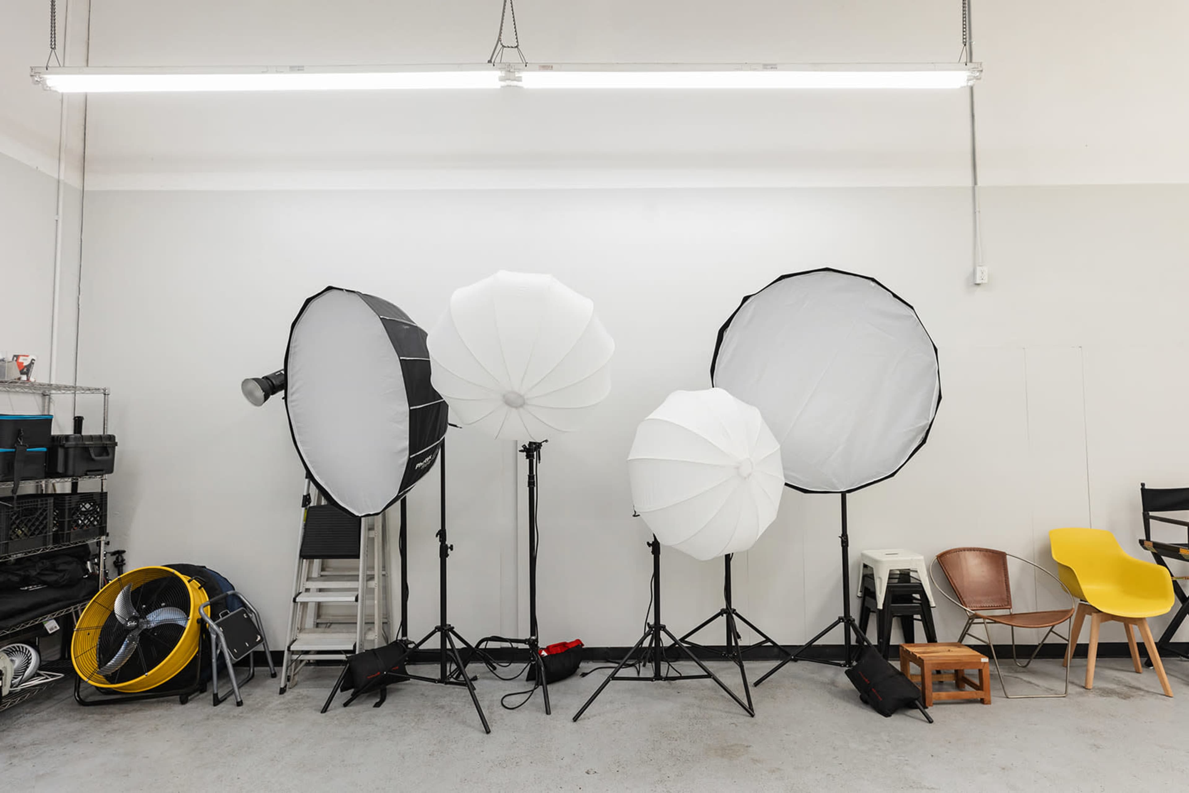 A photography studio with various lighting setups, including three large softboxes on stands and a fan, arranged against a plain wall.