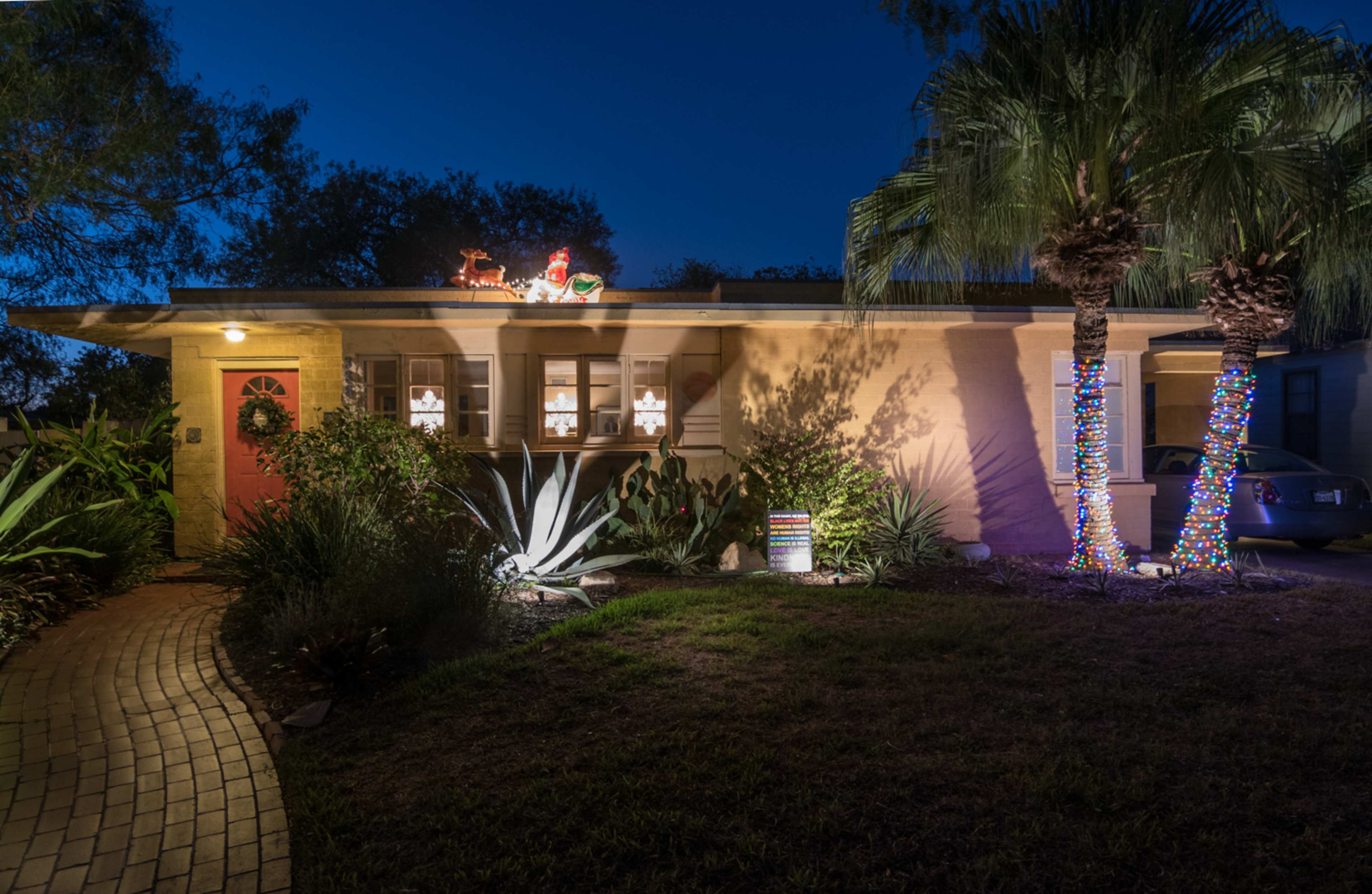 A single-story home is decorated with Christmas lights and a lawn adorned with plants, set against a twilight sky.