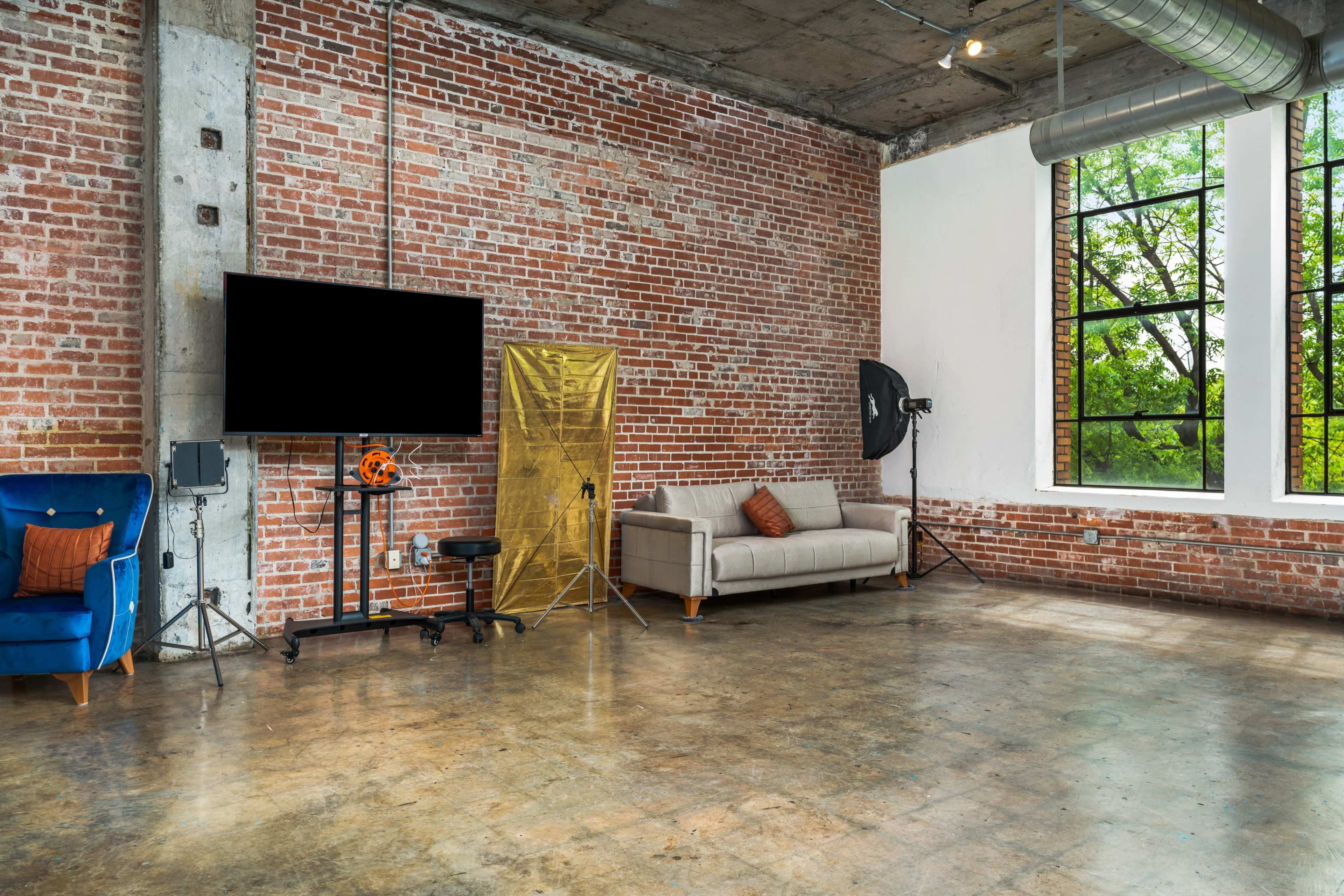 A spacious room with exposed brick walls, a concrete floor, a gray sofa, a television on a stand, and large windows letting in natural light.