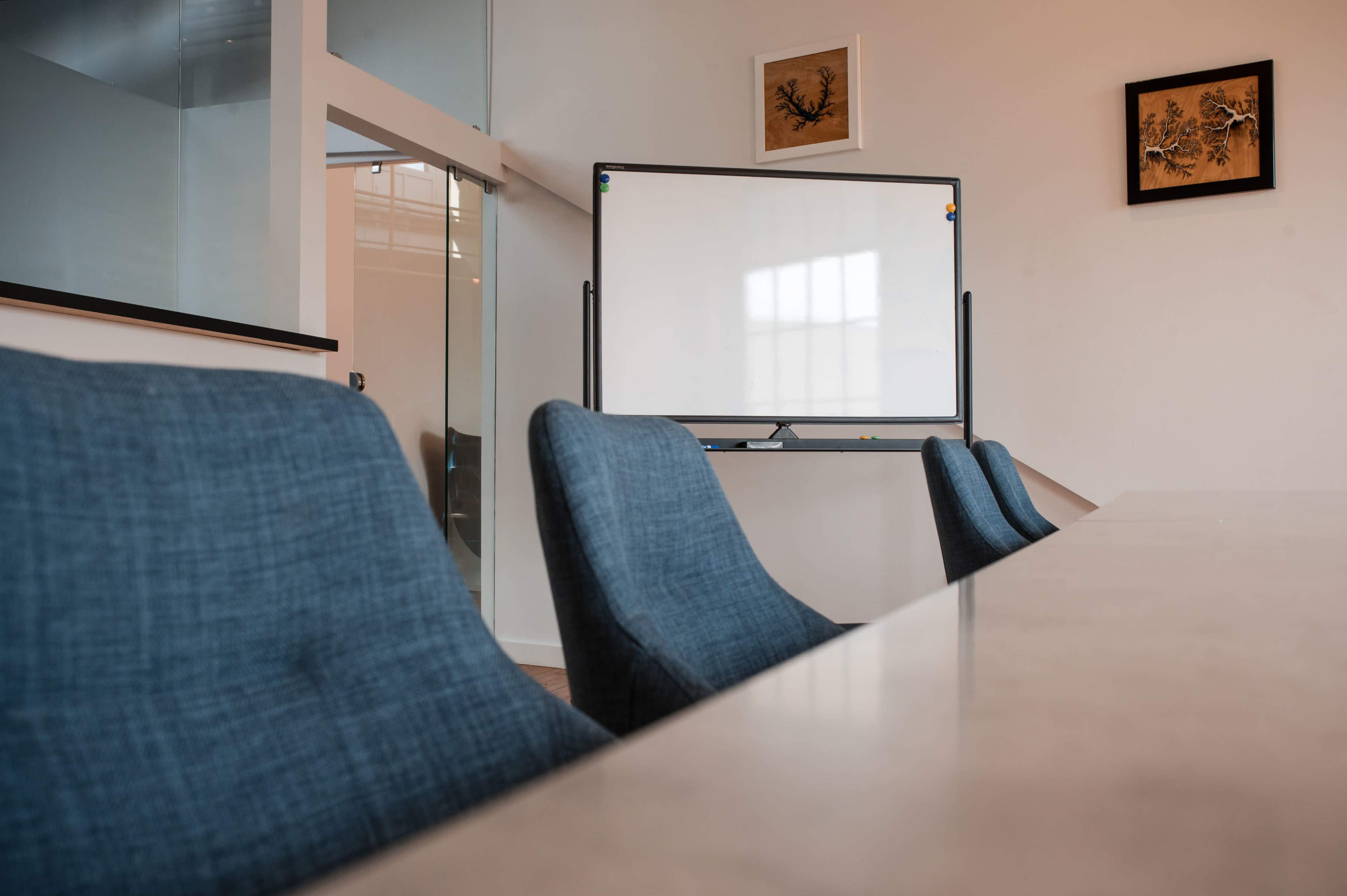 A conference room features a long table with several chairs positioned around it, alongside a whiteboard on a stand against the wall.