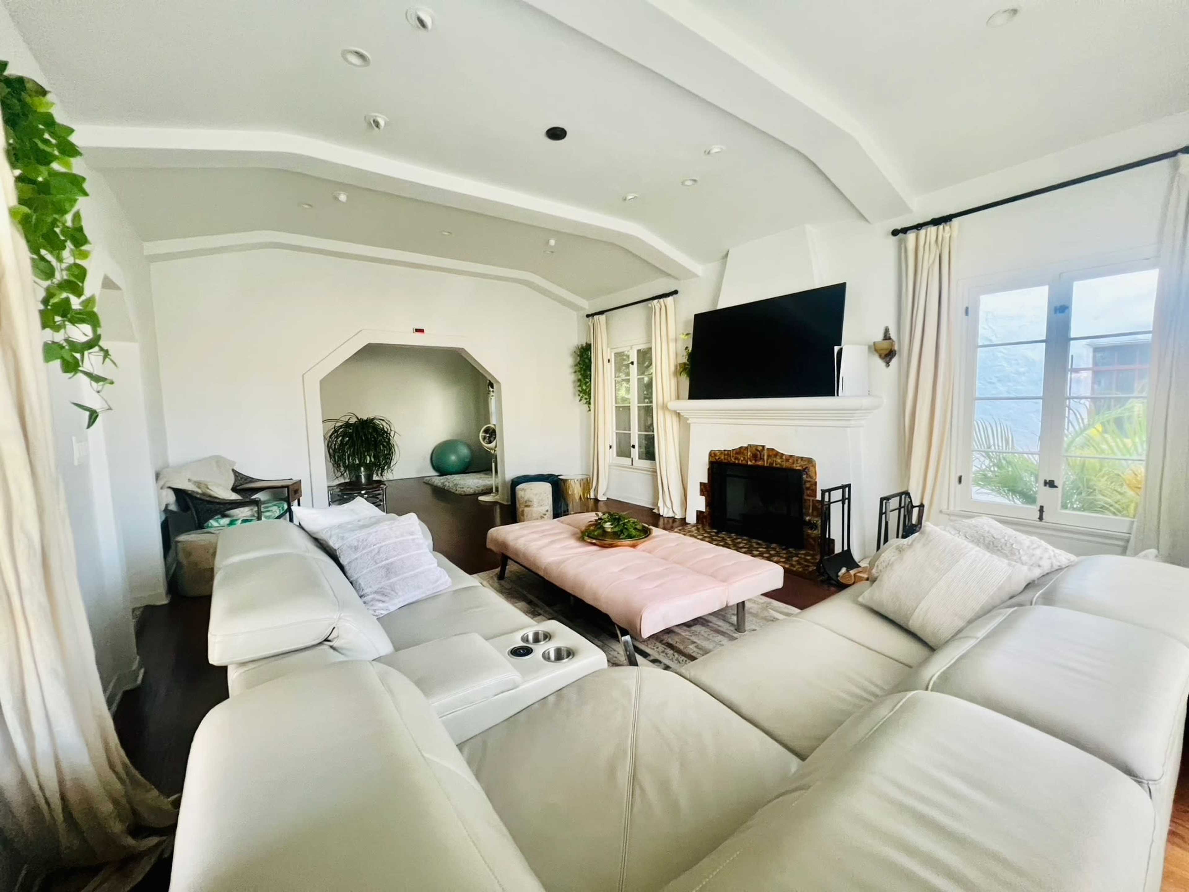 The image shows a modern living room with a light-colored sectional sofa, a pink coffee table, a fireplace, and large windows allowing natural light to enter.