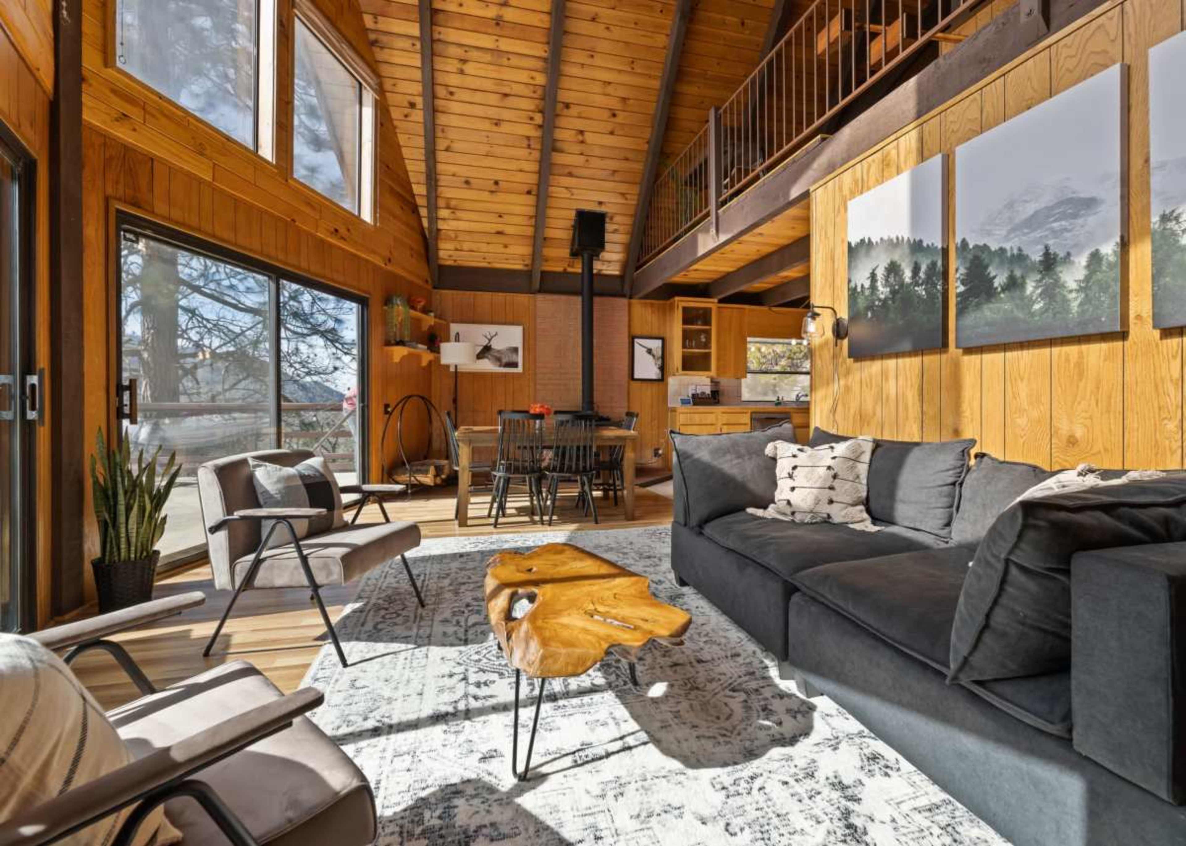A cozy, modern living room with wooden walls and large windows, featuring a couch, a wooden coffee table, and a dining area in the background.