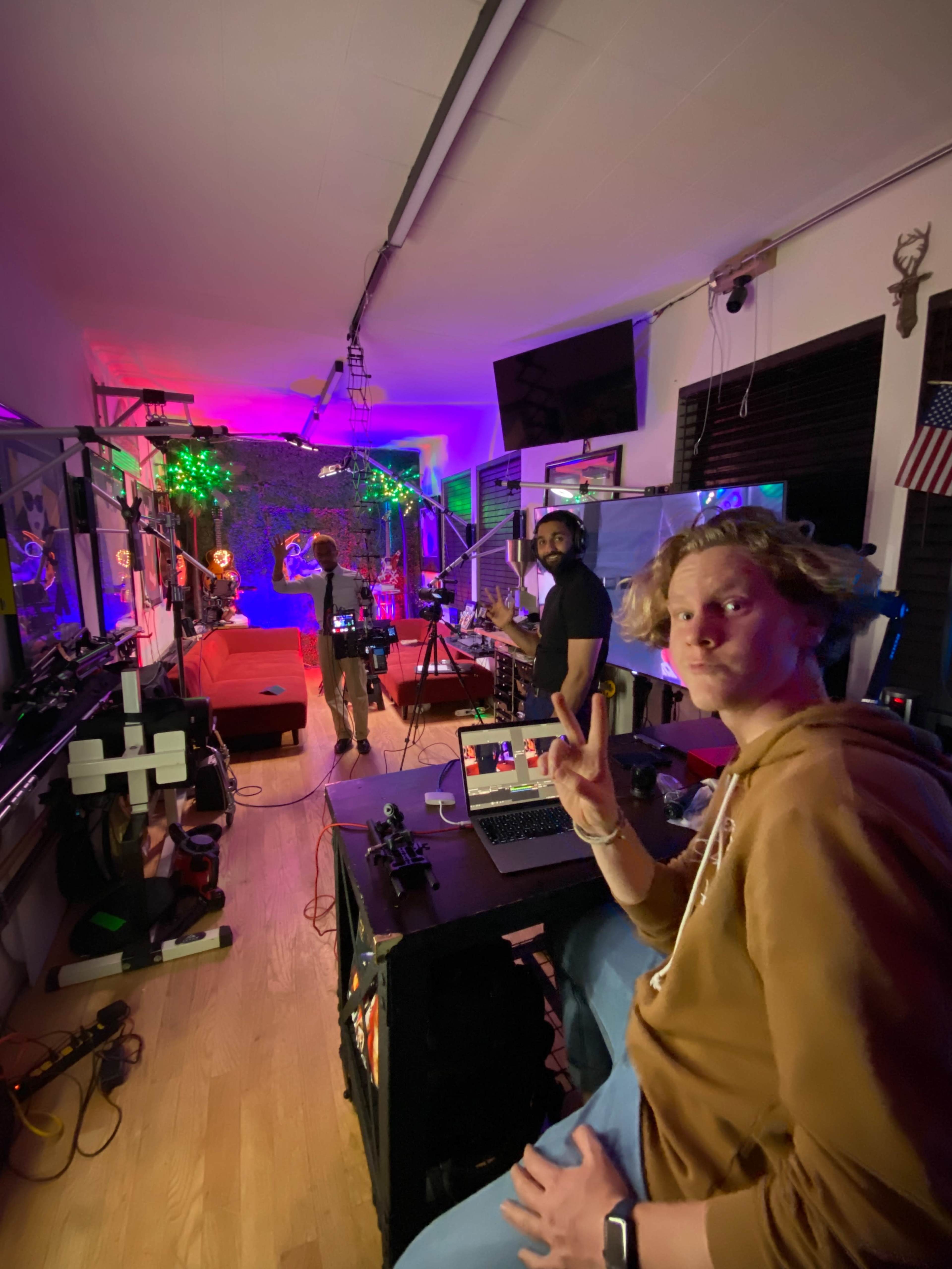 A brightly lit studio space features a person working at a laptop in the foreground, while two others prepare for a shoot in the background surrounded by colorful lighting and equipment.