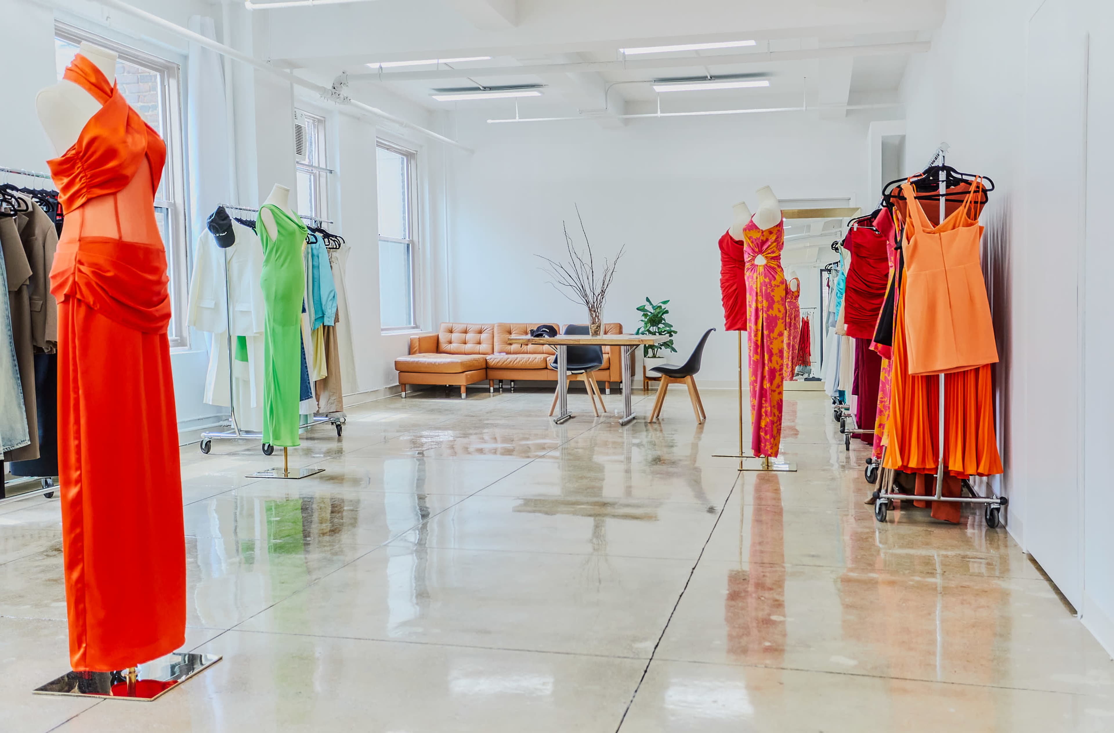 A modern clothing showroom features vibrant dresses displayed on mannequins and racks, with polished concrete floors and a seating area in the background.
