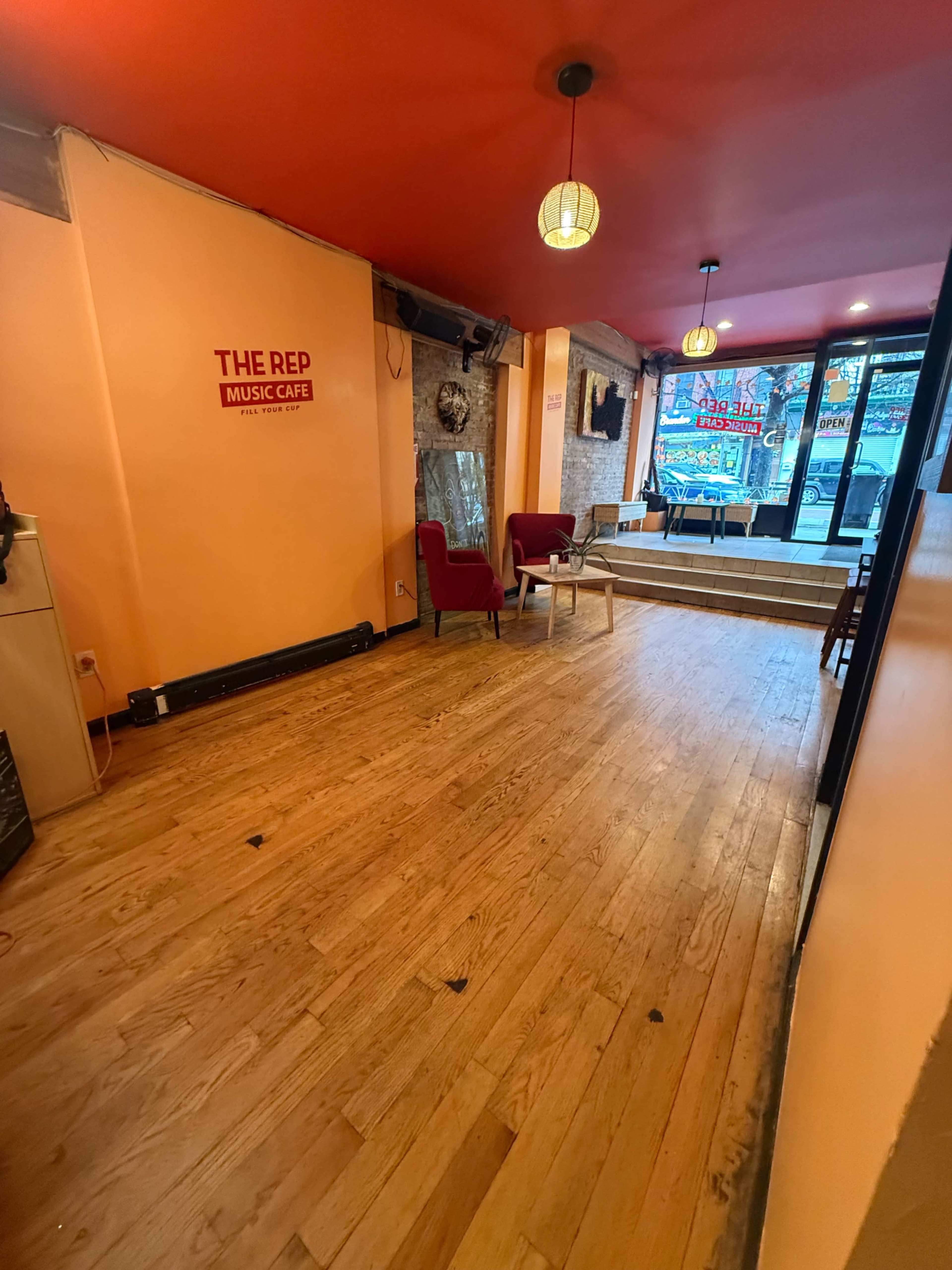 The image shows the interior of a music cafe featuring wooden floors, orange walls, two red chairs, and large windows allowing natural light.
