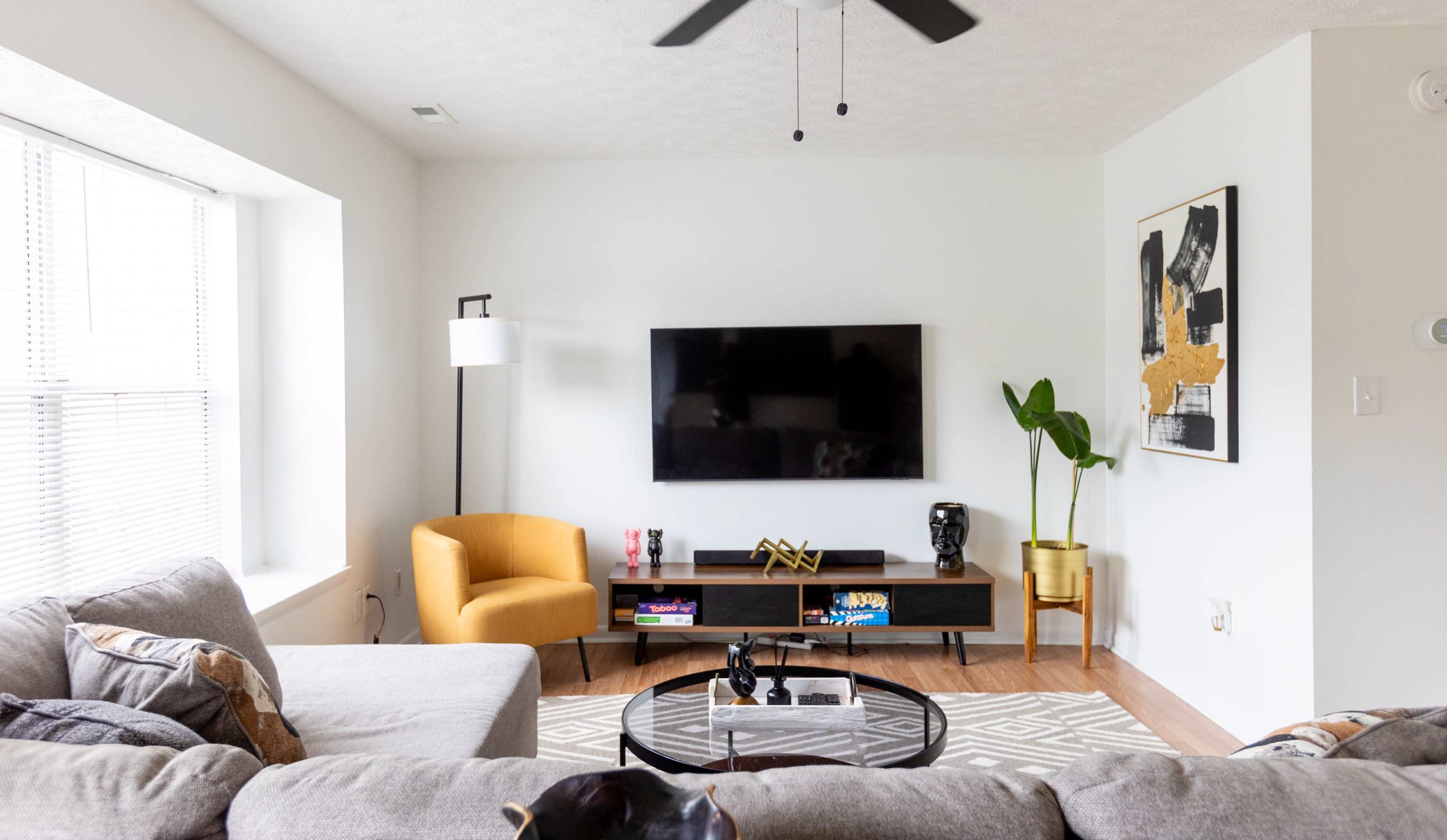 The living room features a gray sectional sofa, a yellow armchair, a black TV mounted on the wall, and a coffee table on a patterned rug.