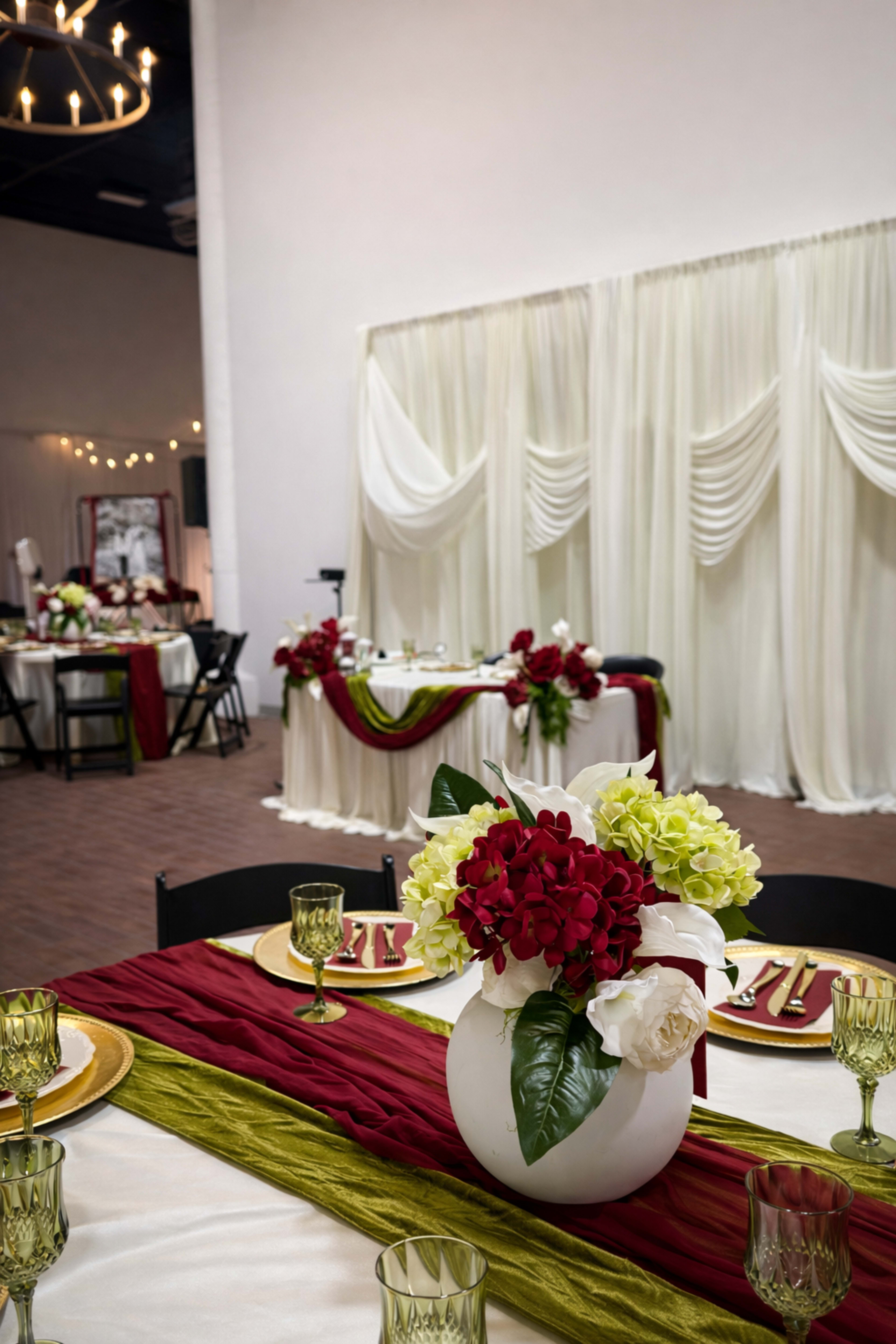 A floral centerpiece featuring white and red flowers is placed on a table adorned with a red and green runner, set in a decorated event space with white drapery and elegant table settings in the background.
