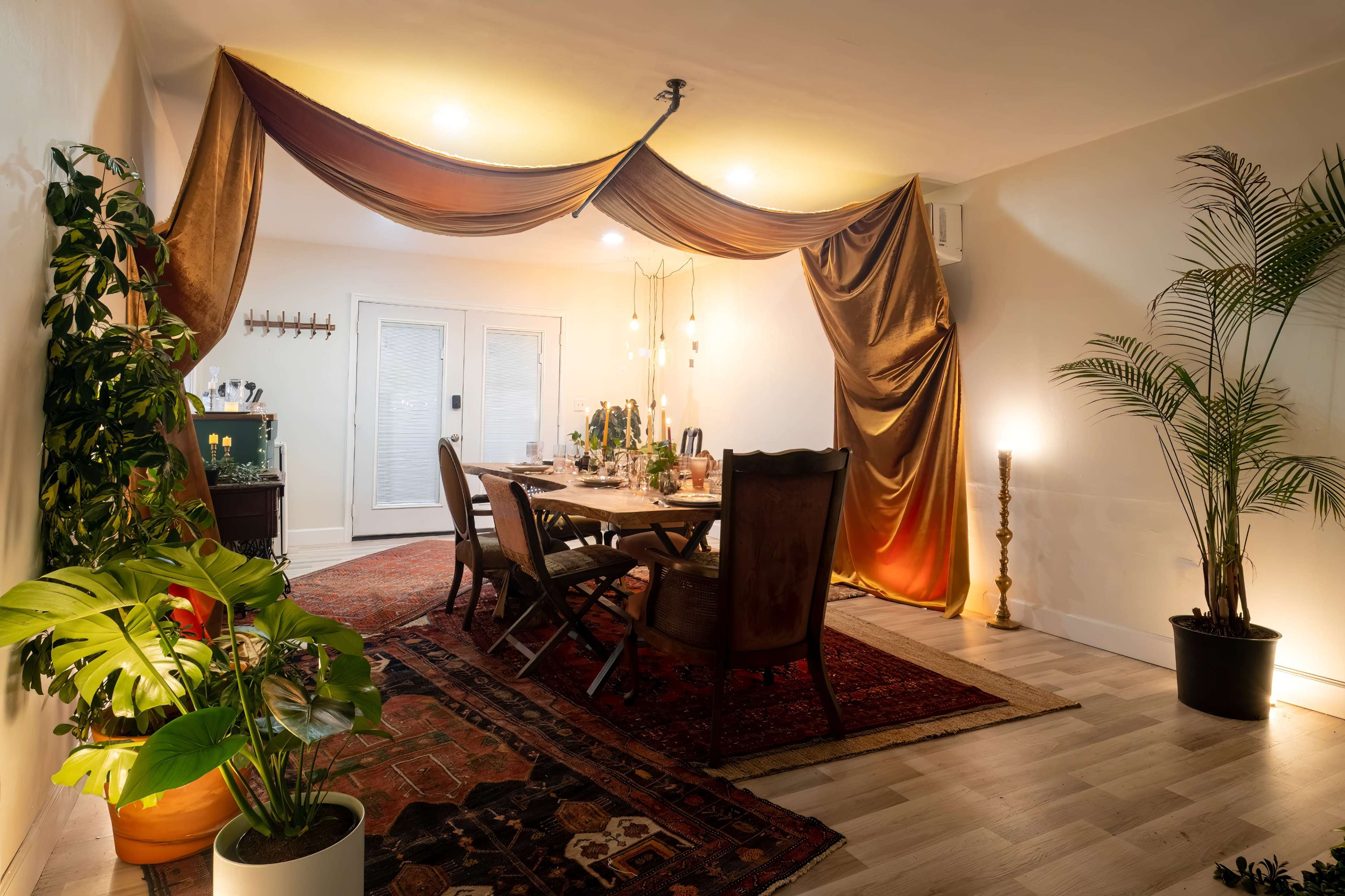 A dining room features a large wooden table surrounded by chairs, illuminated by warm lighting and draped fabric, with plants and a colorful rug accentuating the space.