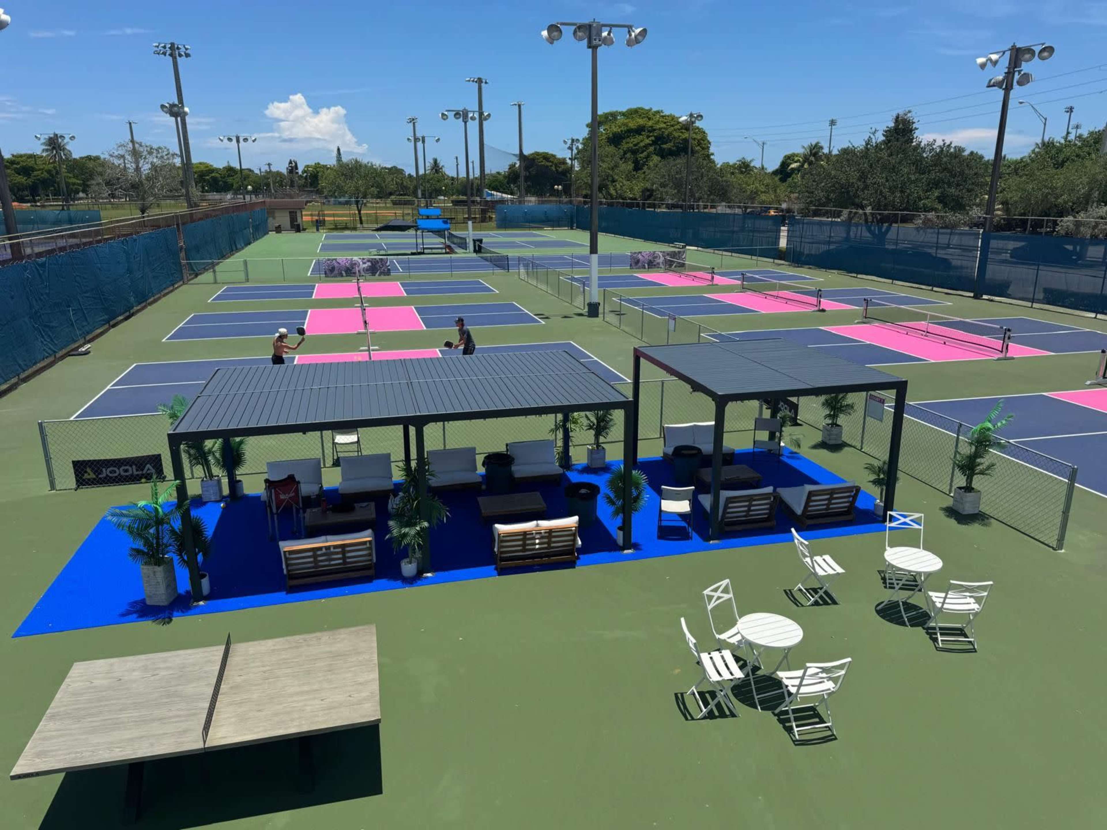 The image shows a tennis facility with multiple courts marked in blue and pink, along with shaded seating areas and tables.