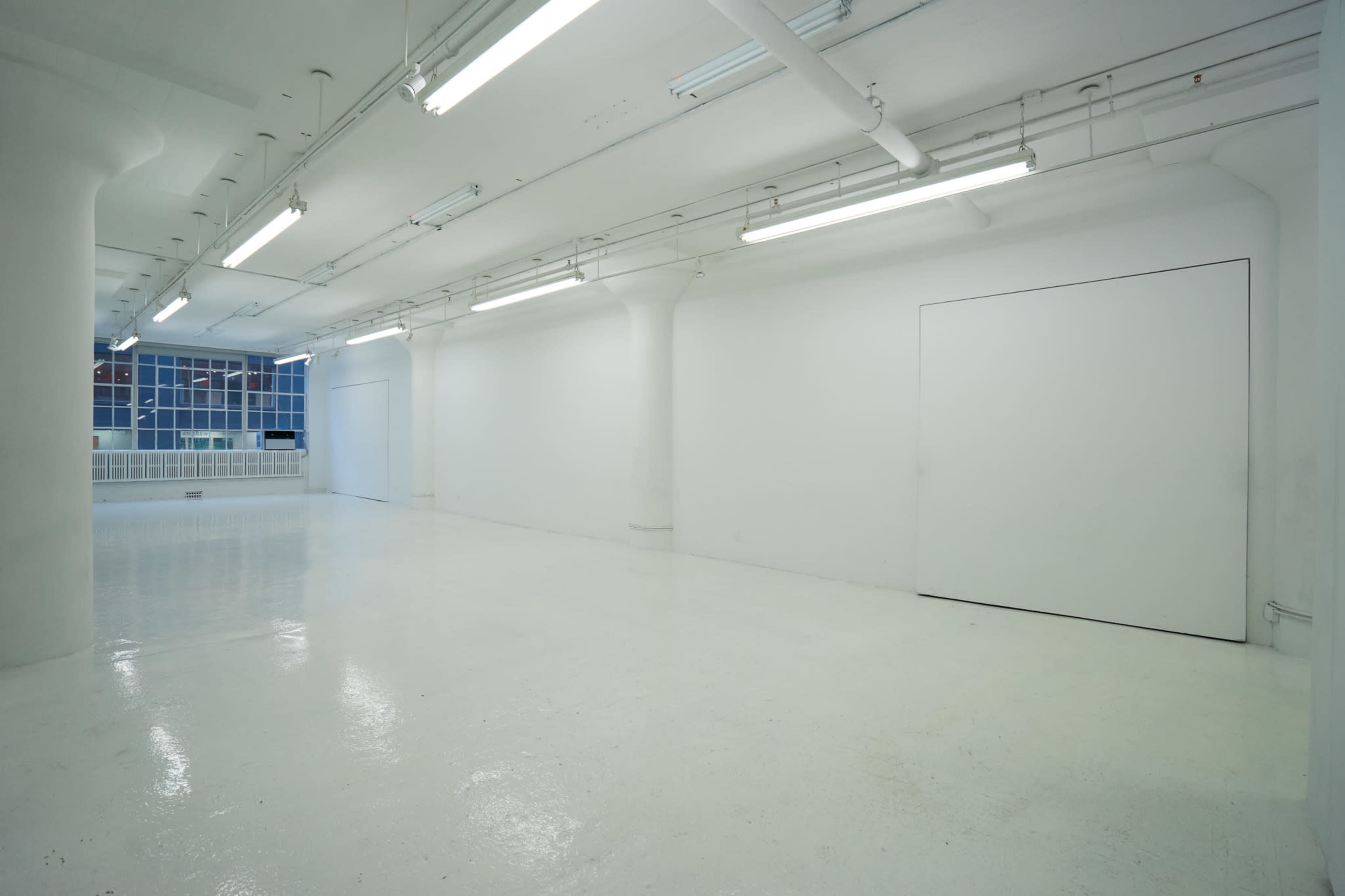 The image shows a spacious, empty room with white walls and a glossy floor, featuring overhead fluorescent lighting and large pillars.