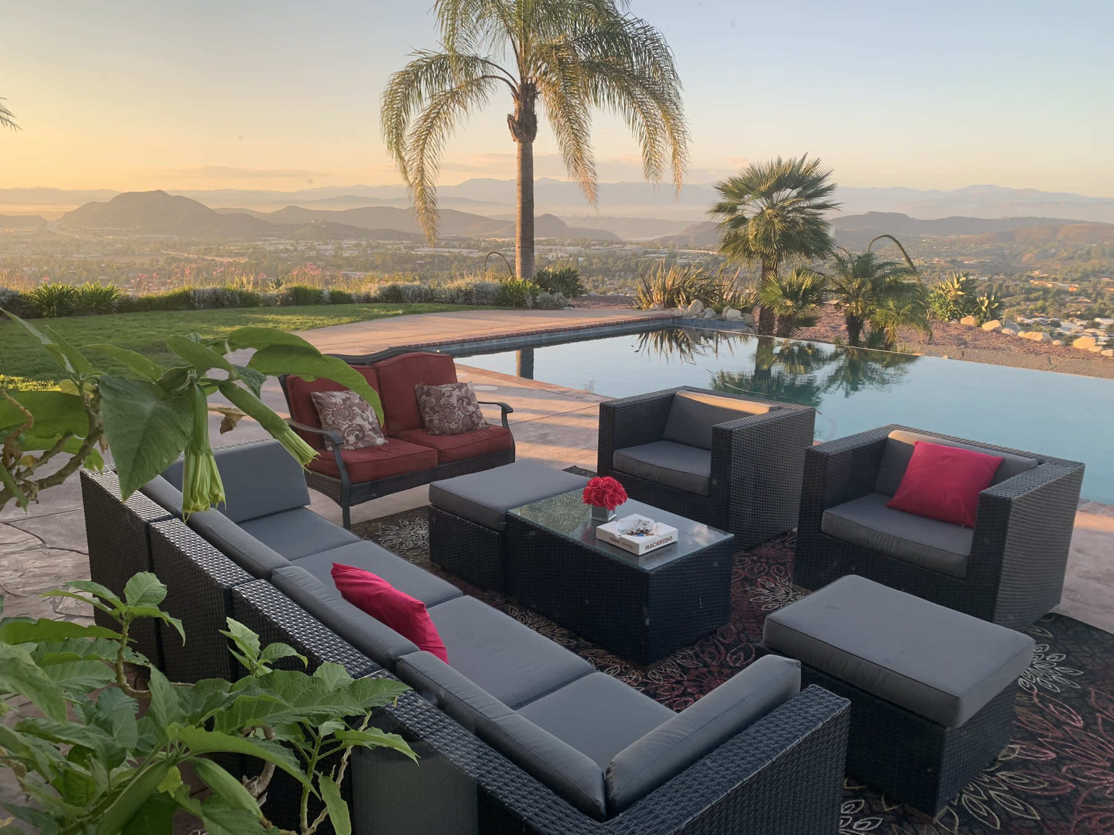 The image shows a modern outdoor seating area with black wicker furniture arranged around a glass table near an infinity pool, overlooking a mountainous landscape at sunset.