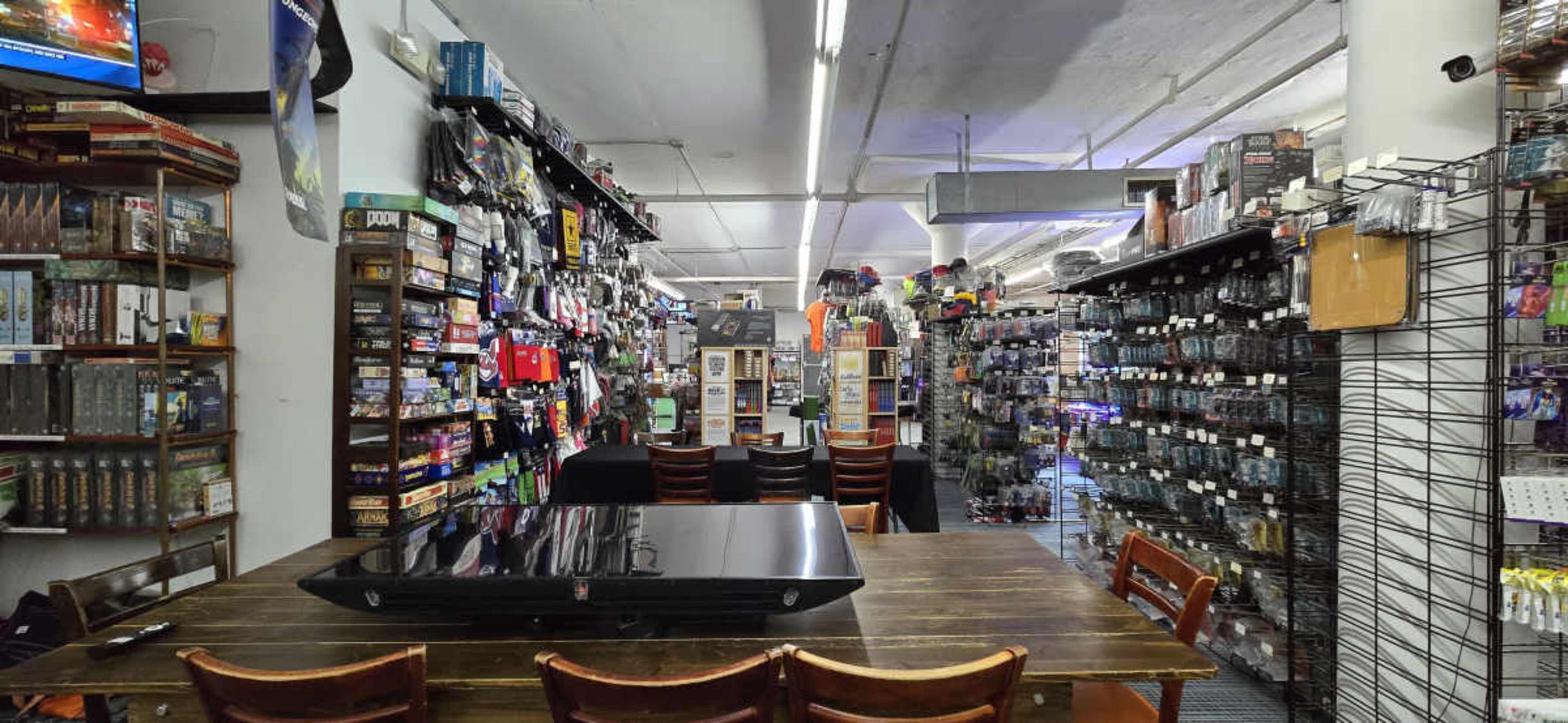 The image shows an organized gaming store with shelves full of board games, collectibles, and gaming accessories, along with a large table in the center for playing games.