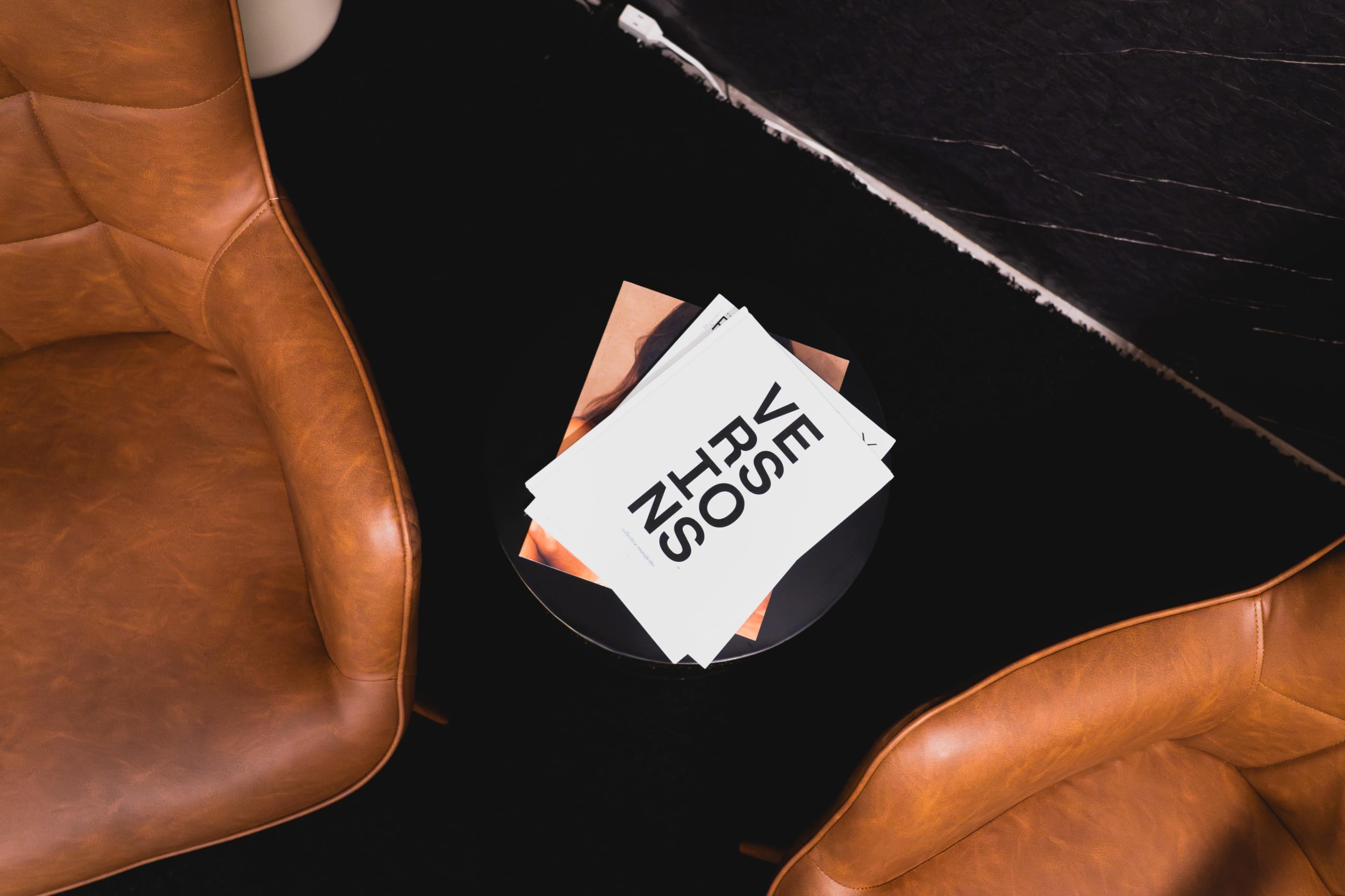 The image features two brown leather chairs positioned beside a black table, which holds a stack of magazines.