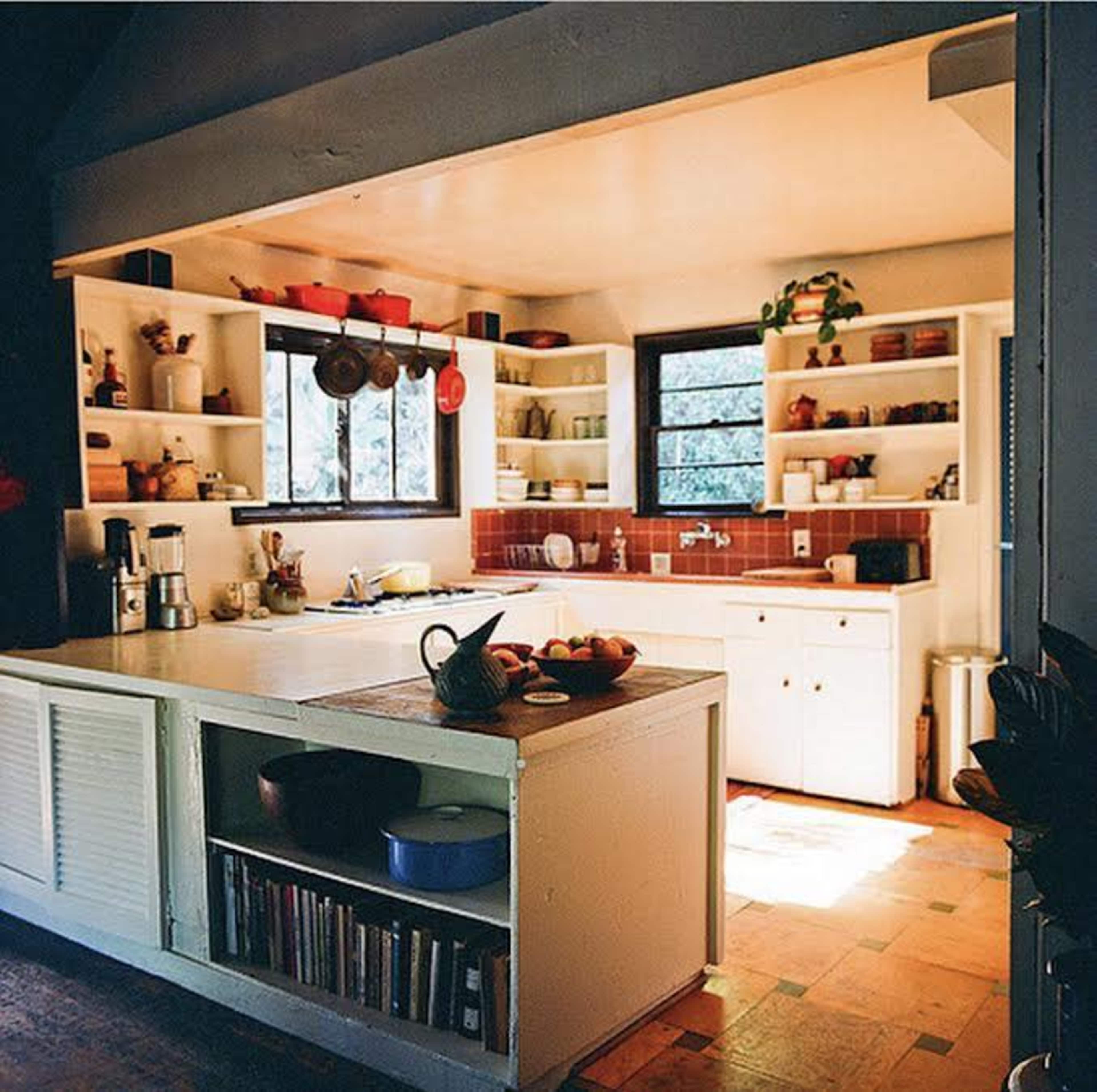 A modest kitchen with open shelves, a central island, and sunlight streaming through a window.