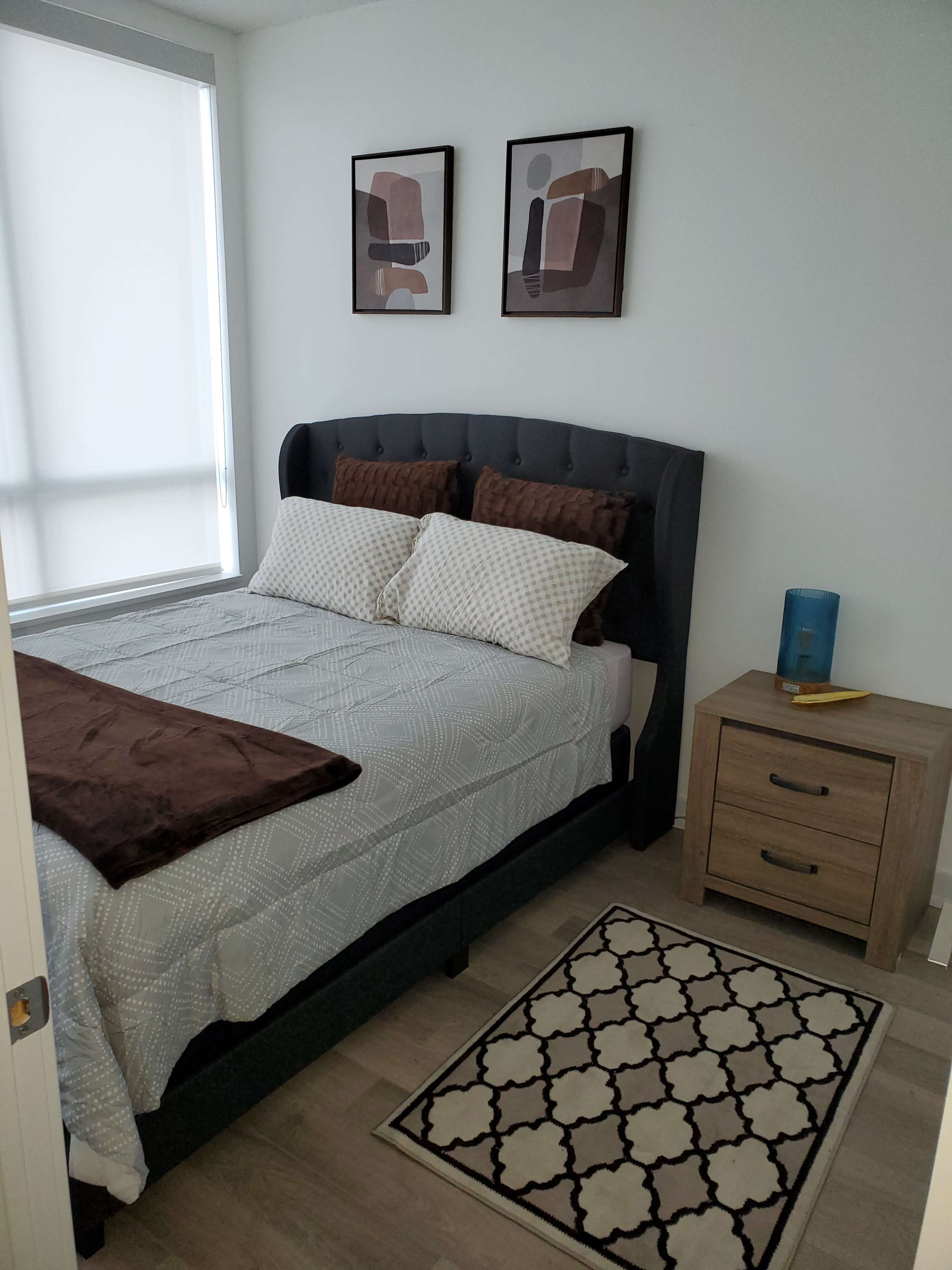 A neatly made bed with decorative pillows in a well-lit bedroom, accompanied by a nightstand and framed artwork on the walls.