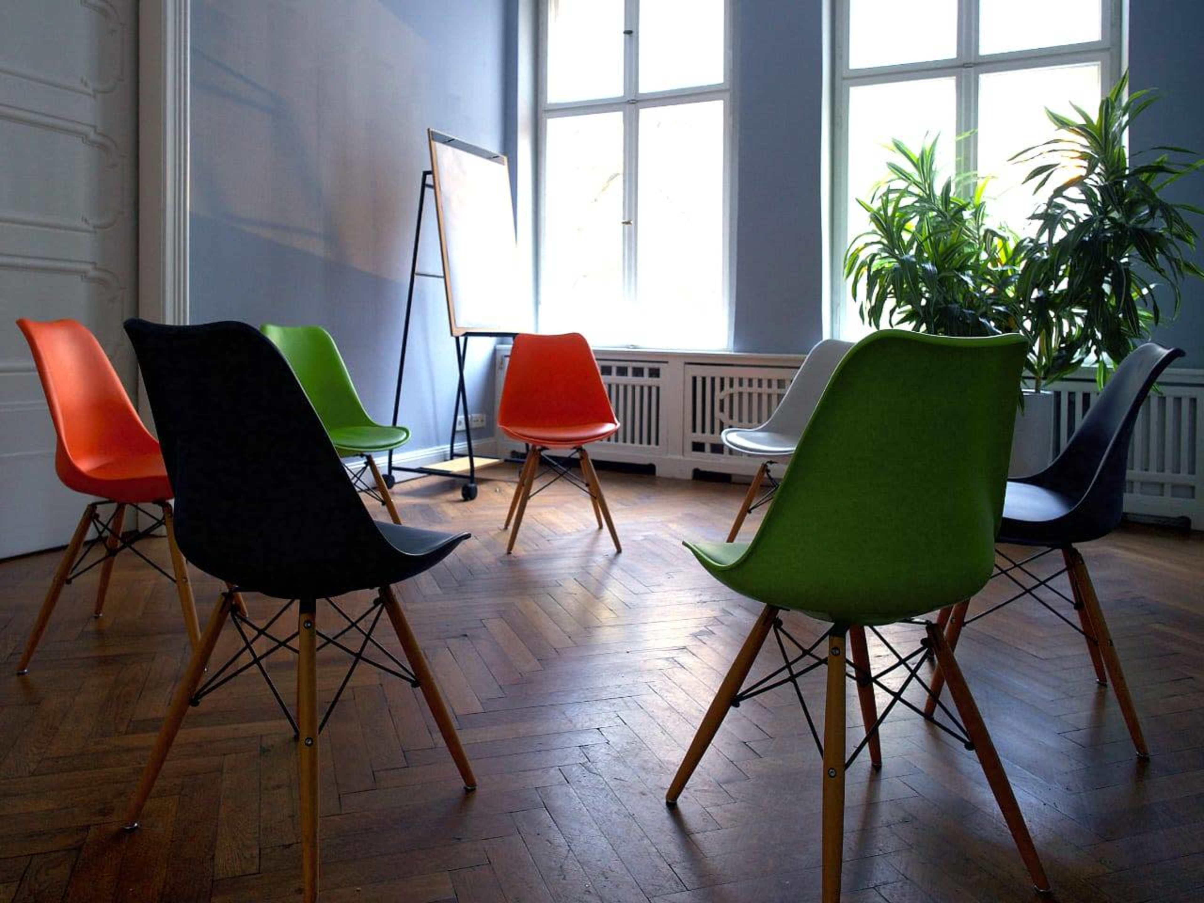 A circle of colorful chairs stands on a hardwood floor in a well-lit room with a large window and a potted plant.