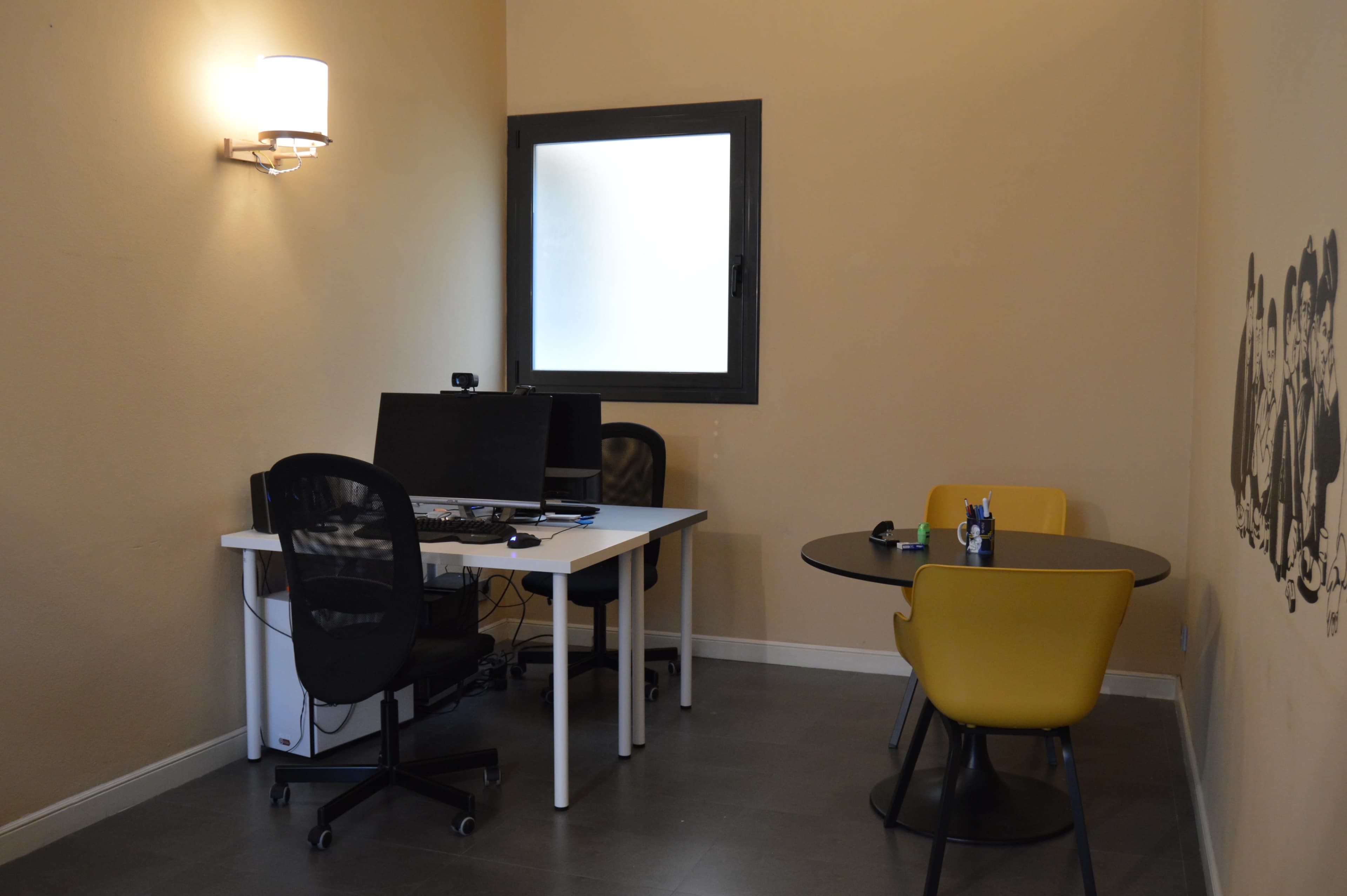 A small office space features two desks, one with monitors, and a round table with chairs.