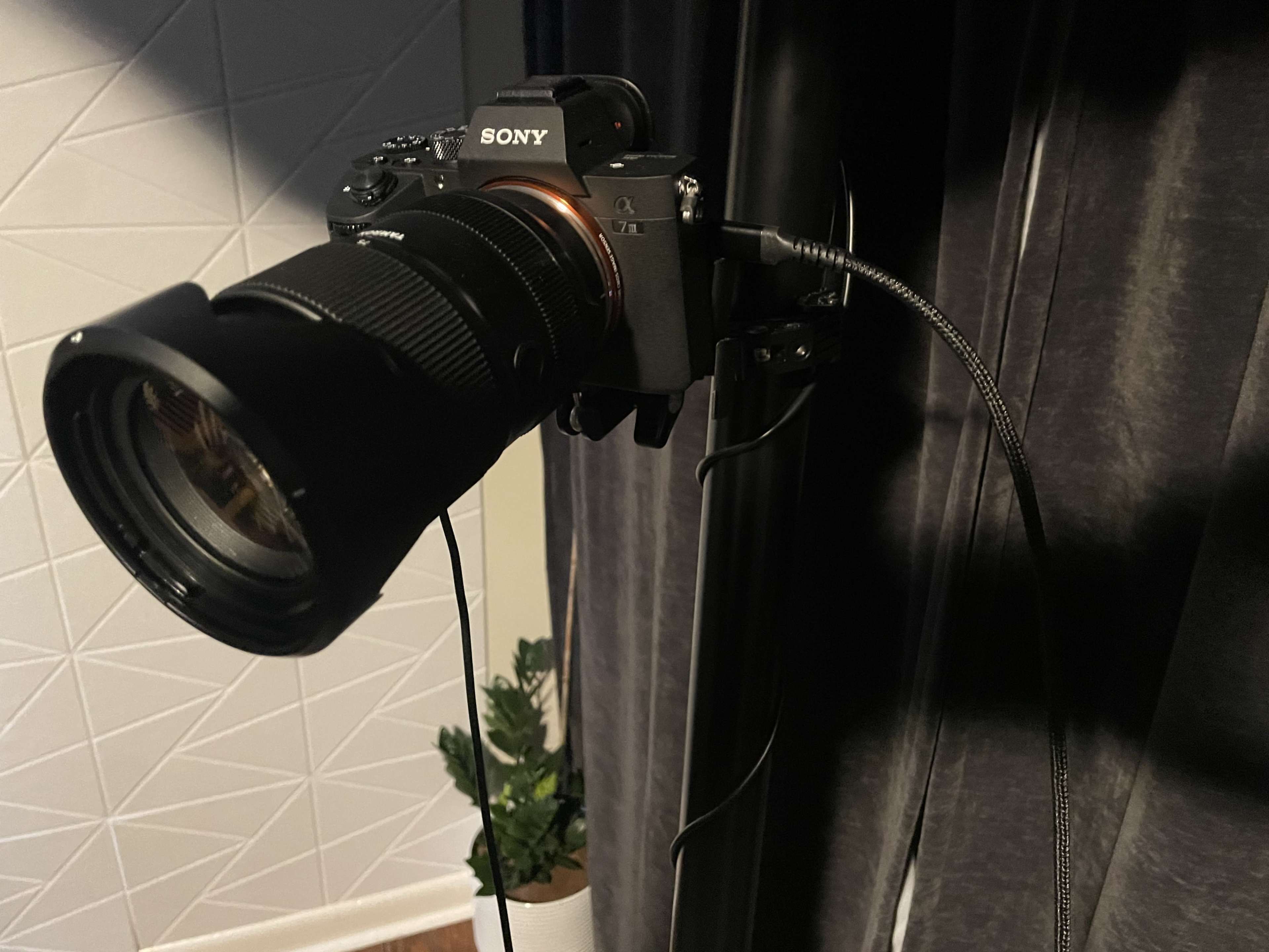 A Sony camera with a large zoom lens is mounted on a stand near a curtain, with a cable connected to it.
