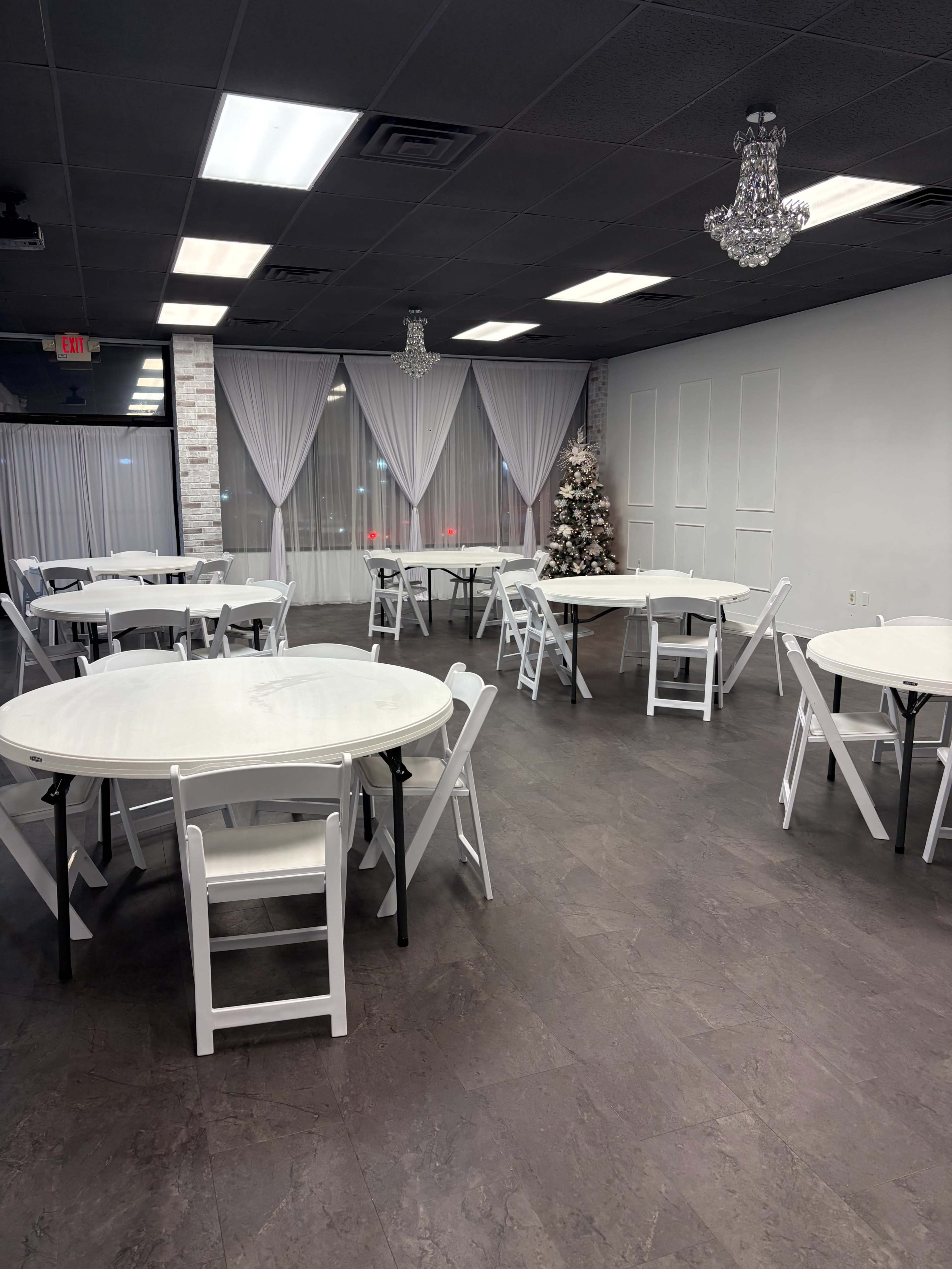 The image shows a spacious event room with several round tables and white folding chairs, decorated simply with a Christmas tree and sheer drapes.