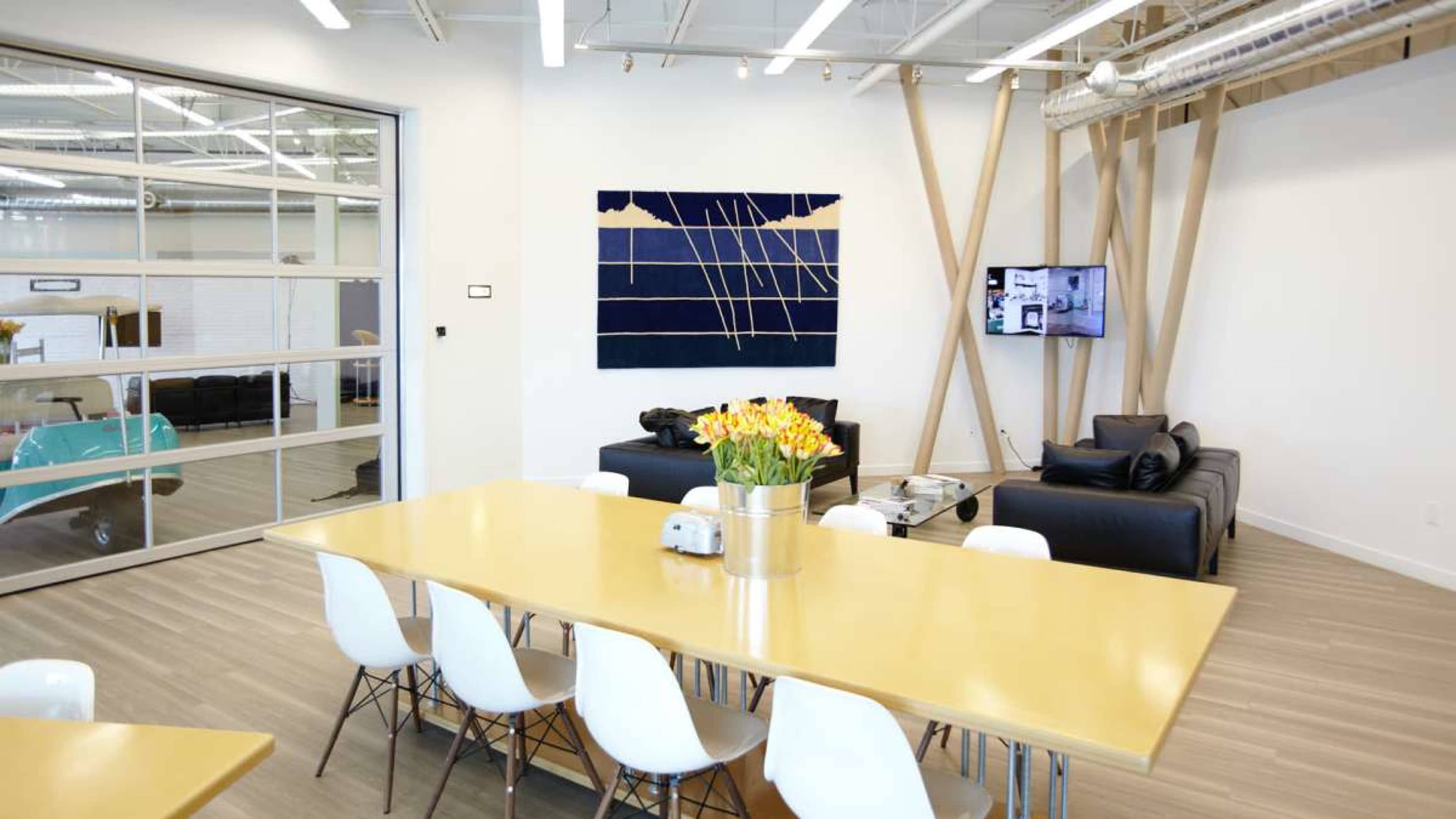 The image shows a modern office space with a long yellow table surrounded by white chairs, black sofas in the background, and an abstract wall hanging above the table.