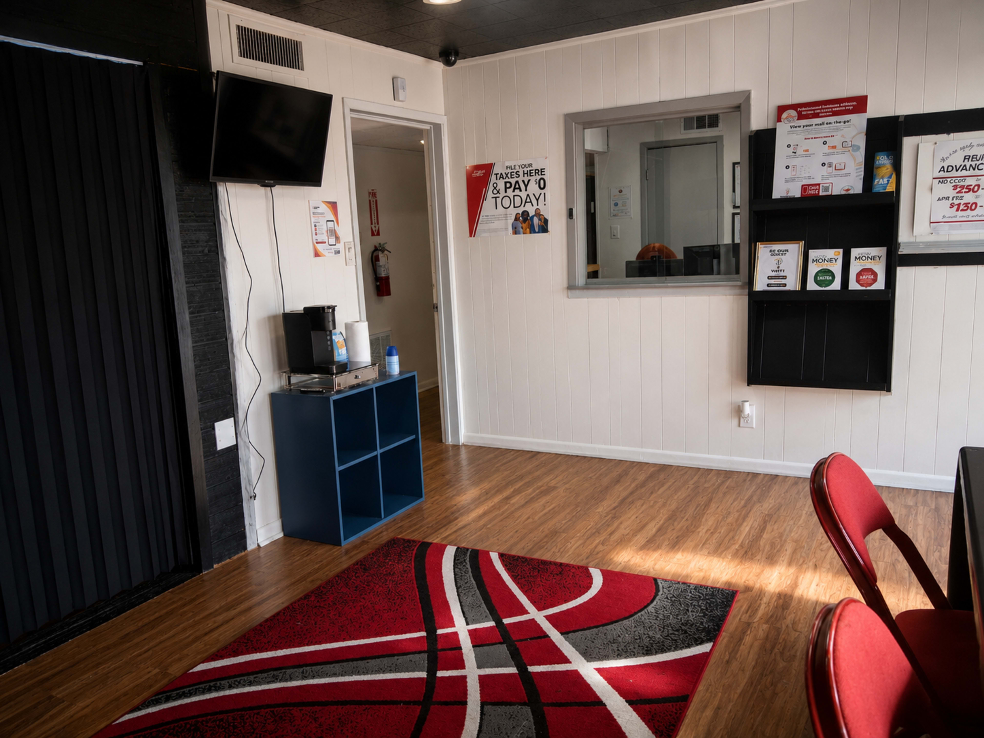 The image shows a small room with a coffee station, a television mounted on the wall, a reception area with a glass counter, and red chairs arranged around a table on a patterned rug.