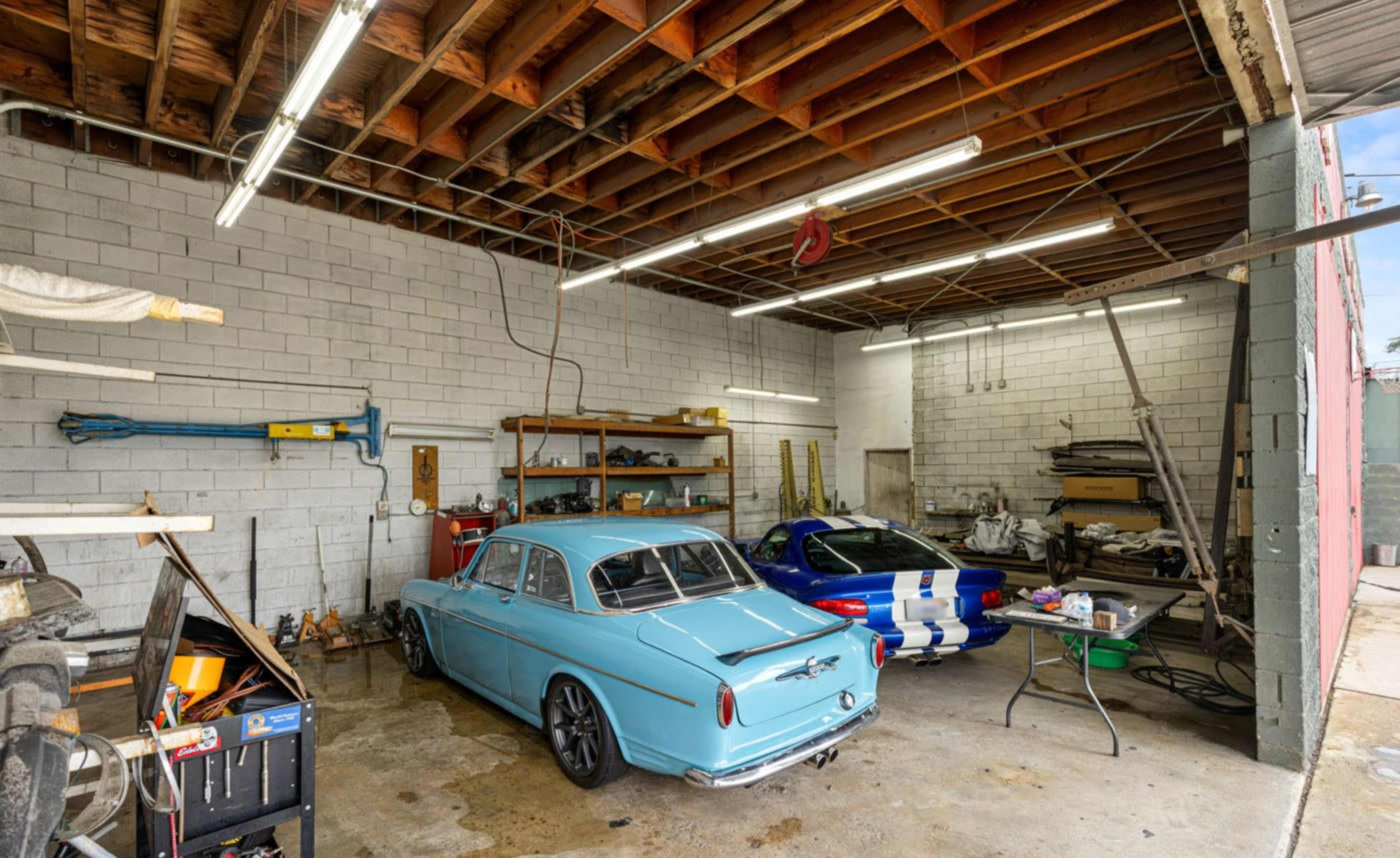 A garage interior with two cars, a light blue sedan and a blue sports car, along with various tools and equipment arranged in the space.