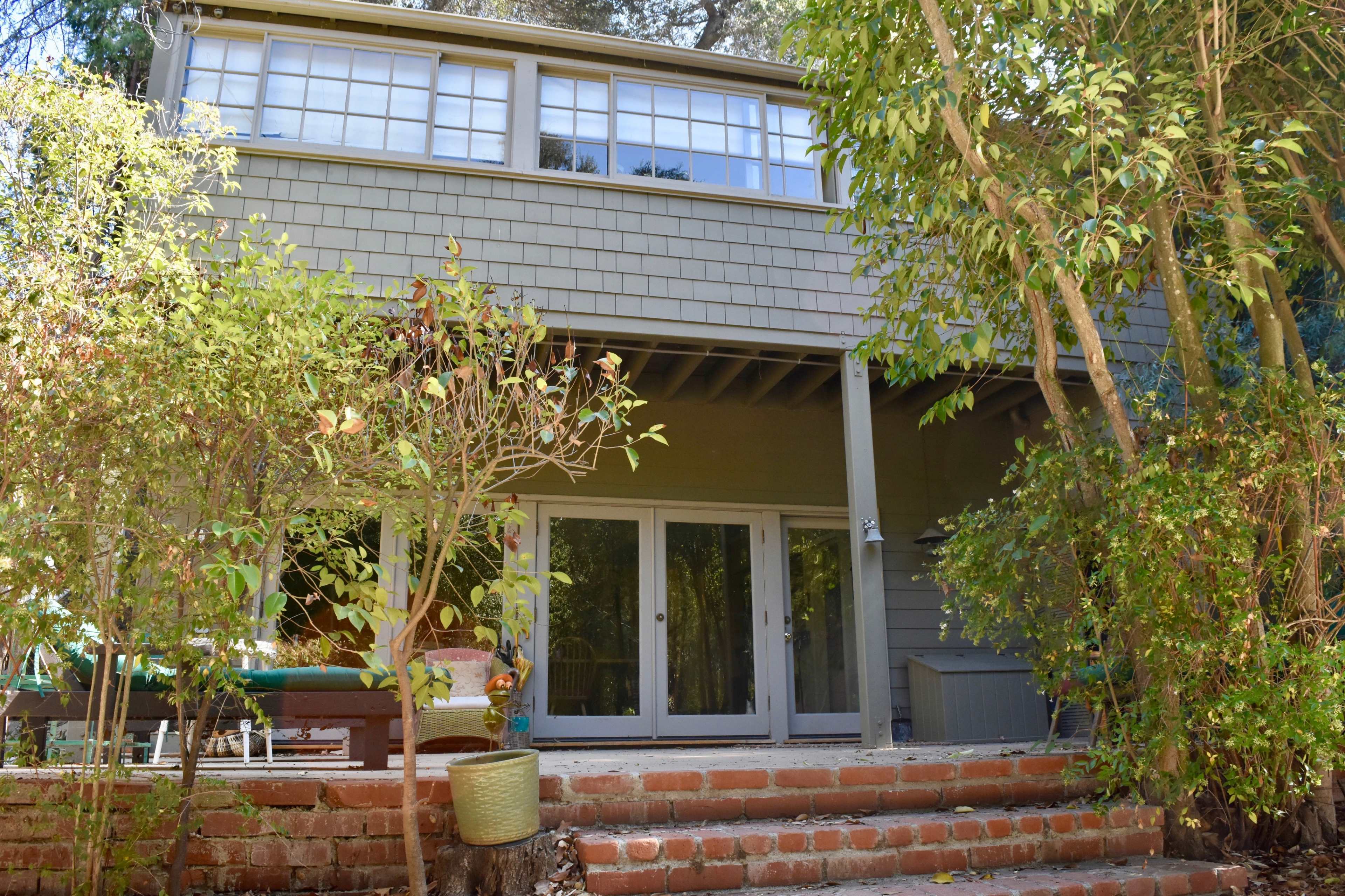 A two-story house with large windows is set among greenery and features a brick staircase leading to a patio area.