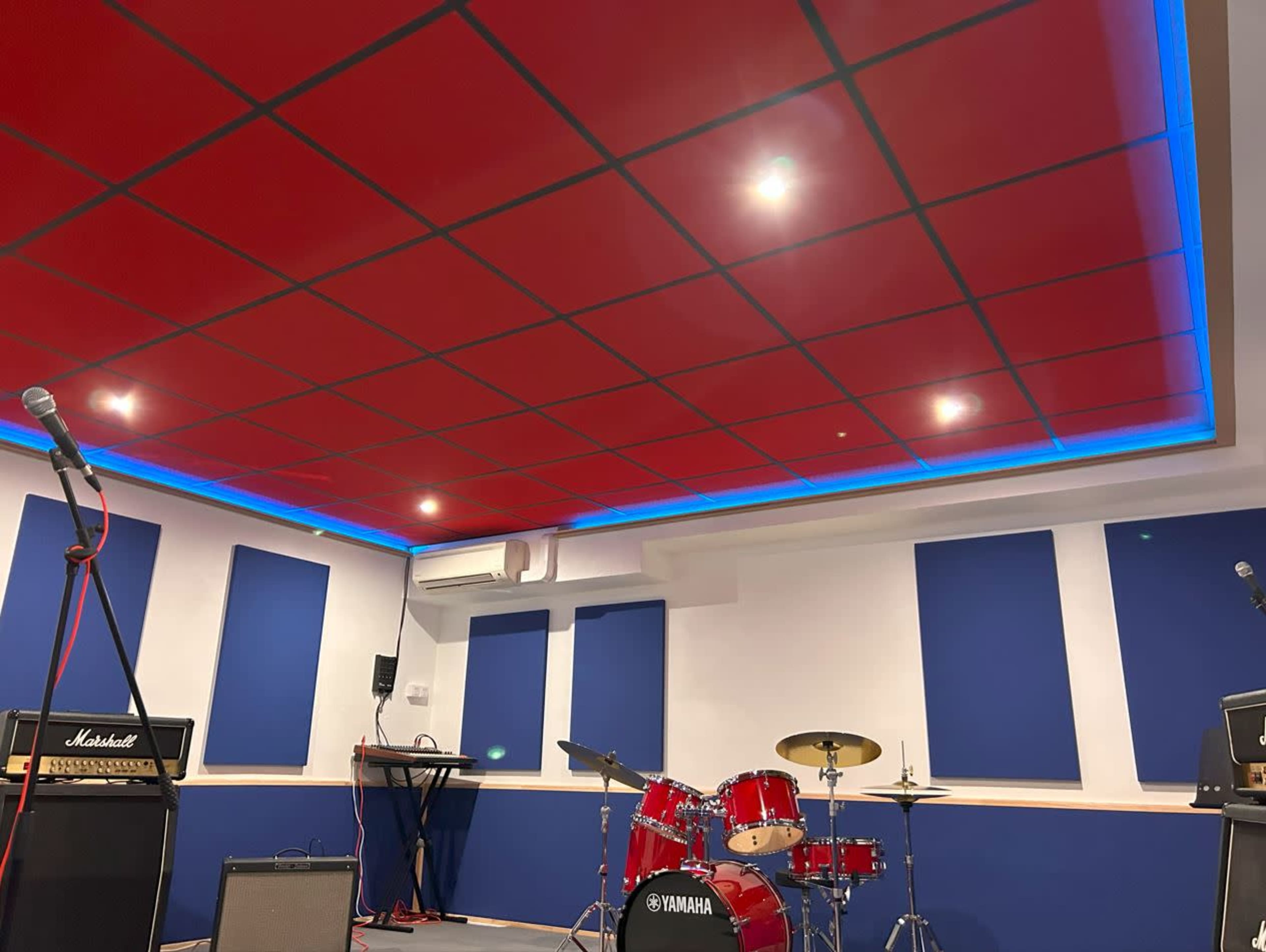 A music rehearsal studio featuring a red ceiling, blue soundproofing panels on the walls, a drum set, and amplifiers.