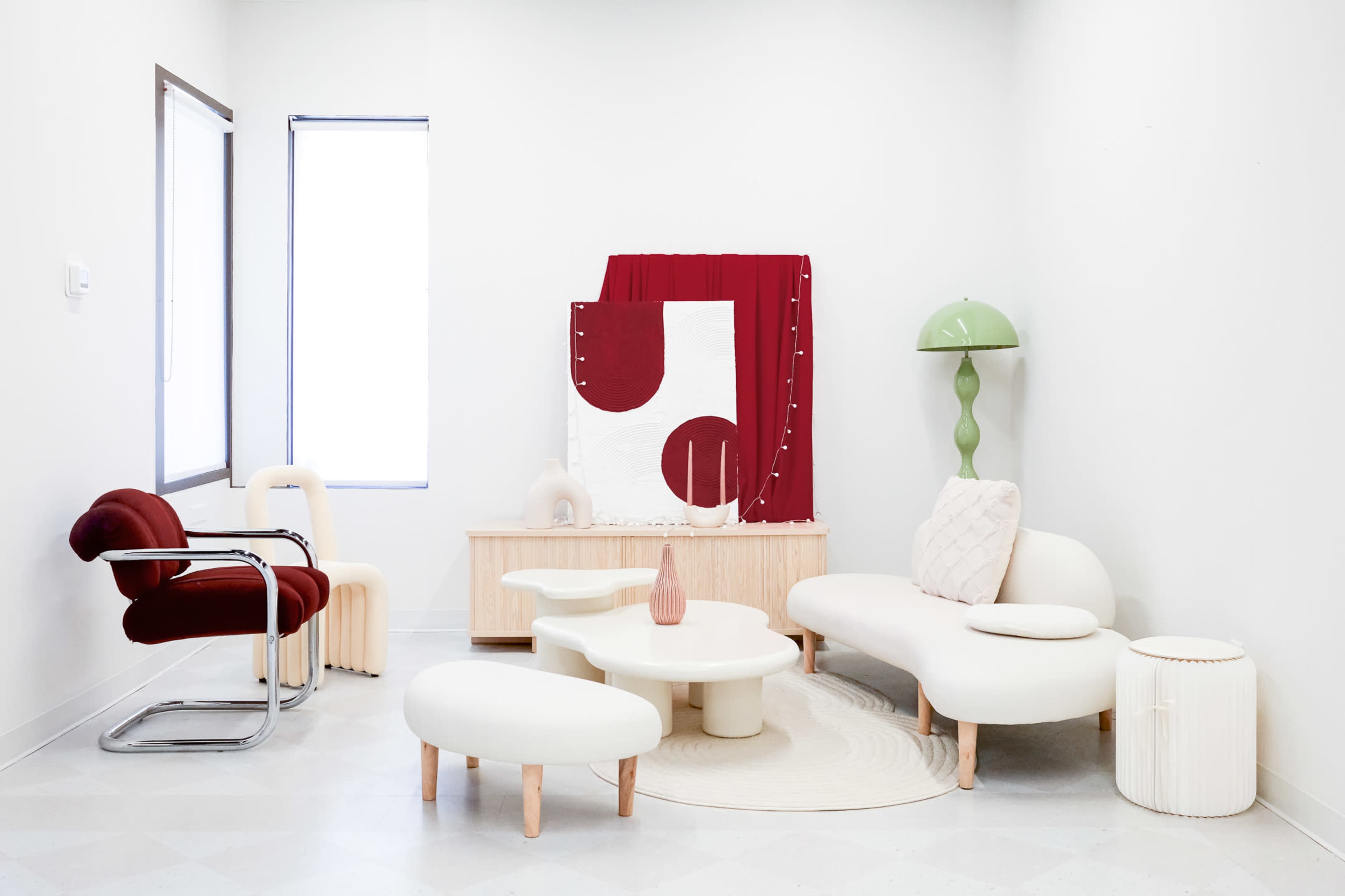 A modern, minimalist living space featuring a curved white sofa, a round coffee table, a red accent chair, and decorative elements like a green lamp and a red wall hanging.