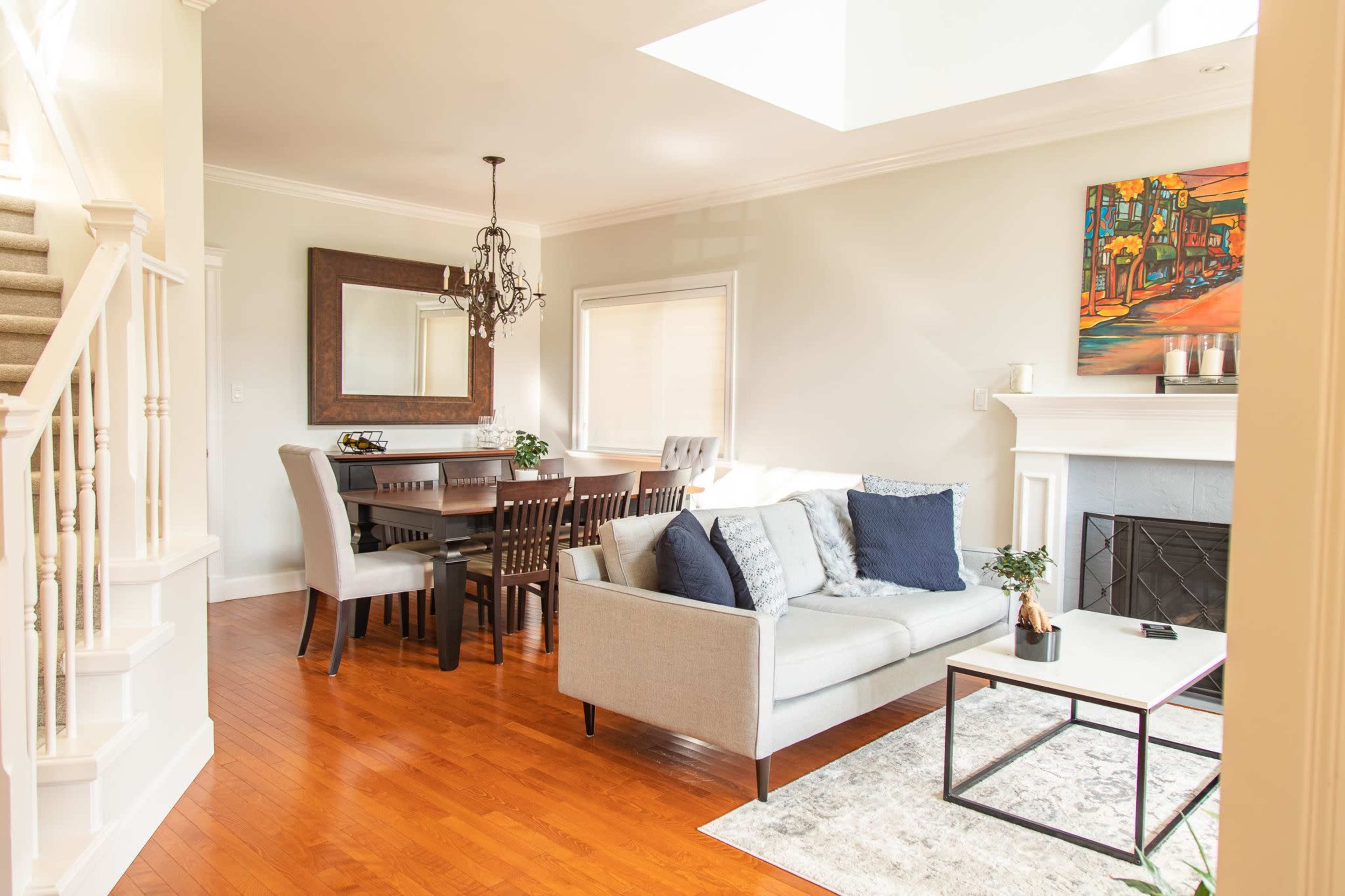 A bright living and dining area with a gray sofa, a wooden dining table, and a fireplace beneath a colorful painting.