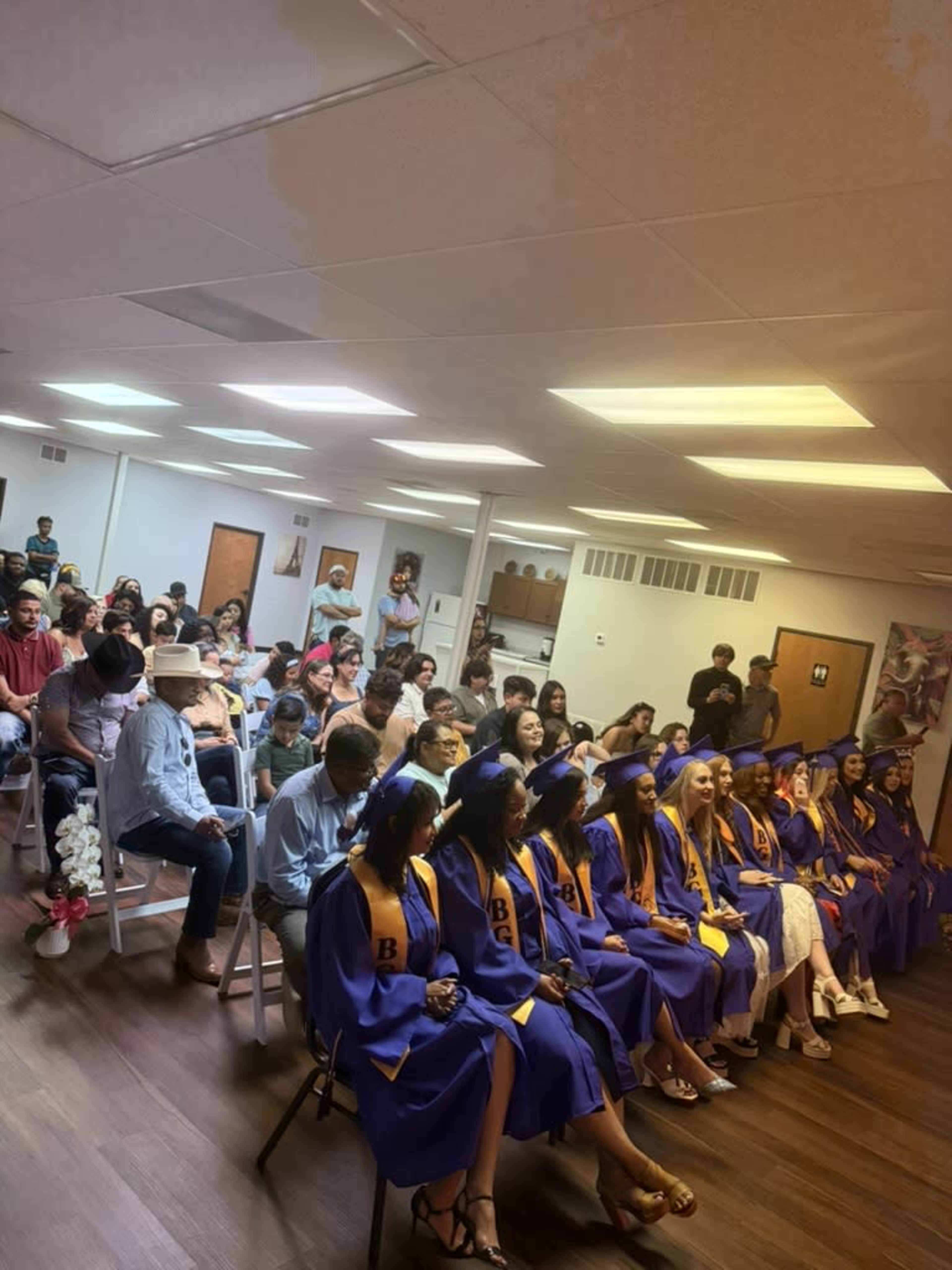 A group of graduates in purple caps and gowns sits in rows, while family and friends fill the seats in a brightly lit indoor venue.