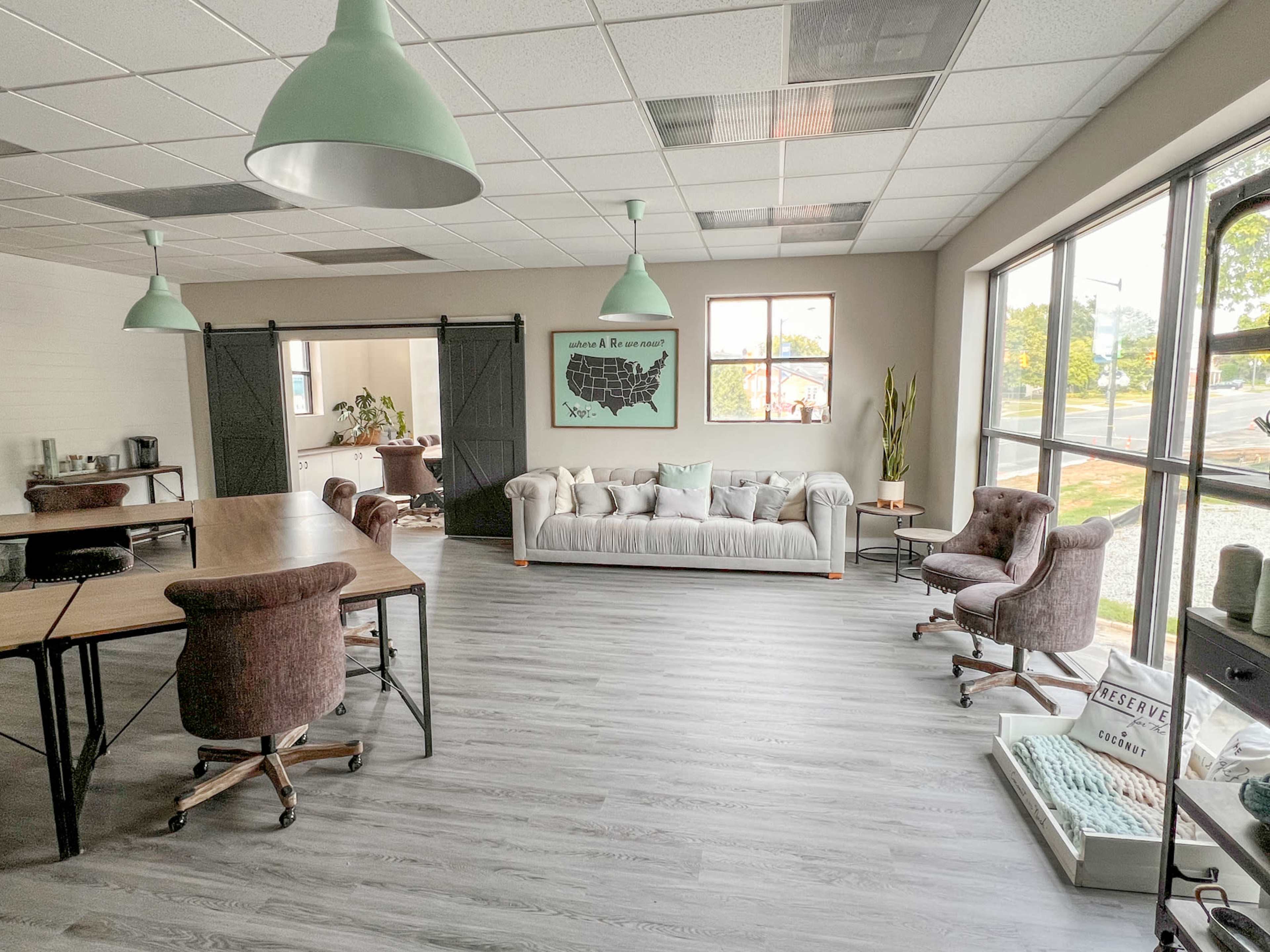 The image shows a spacious, modern office environment featuring a light-colored sofa, a large table with chairs, and large windows allowing natural light to enter.