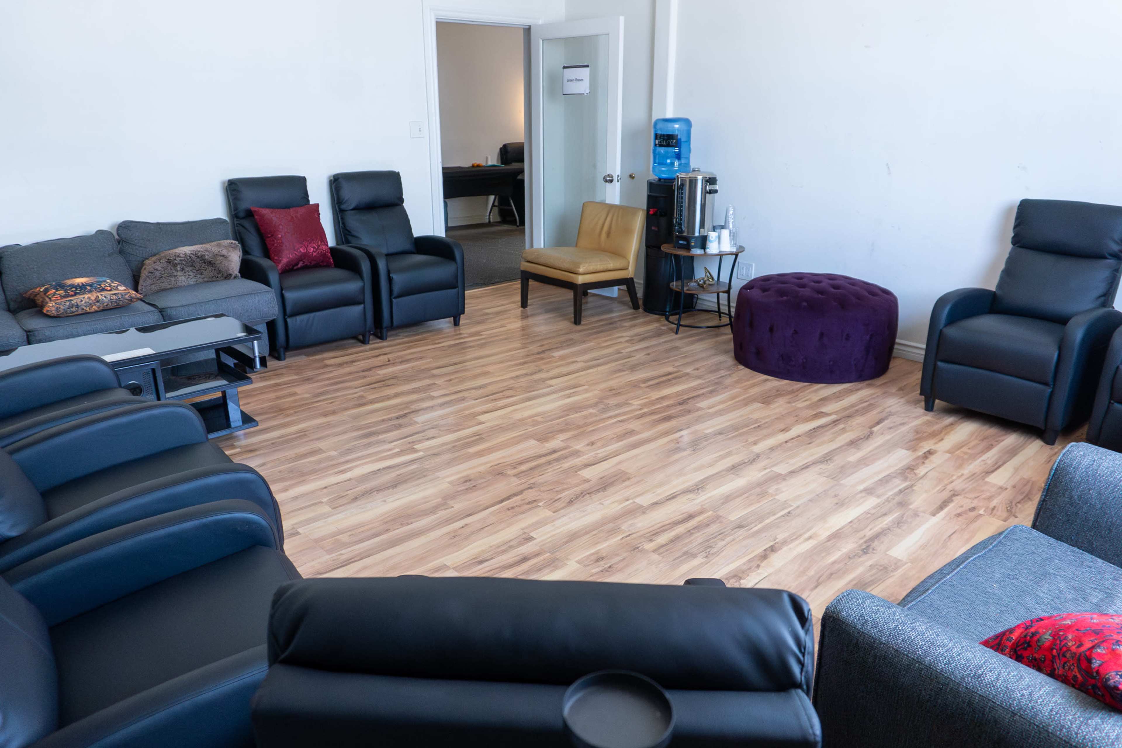 The image shows a seating area with several recliner chairs, a small round ottoman, and a water cooler, arranged in a circular formation on a wooden floor.