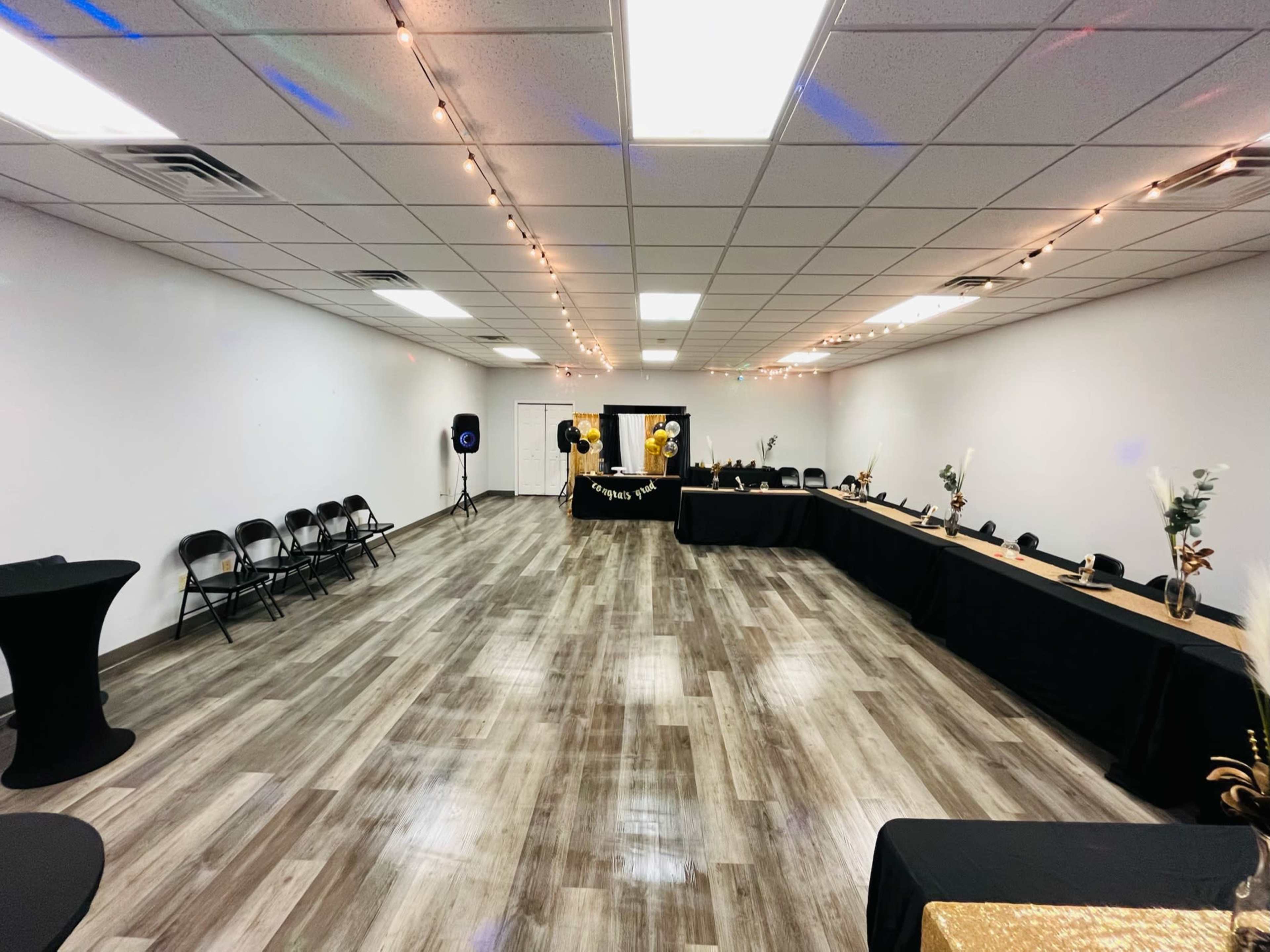 A spacious event room with a row of black chairs and tables decorated in black and gold, surrounded by string lights on the ceiling.