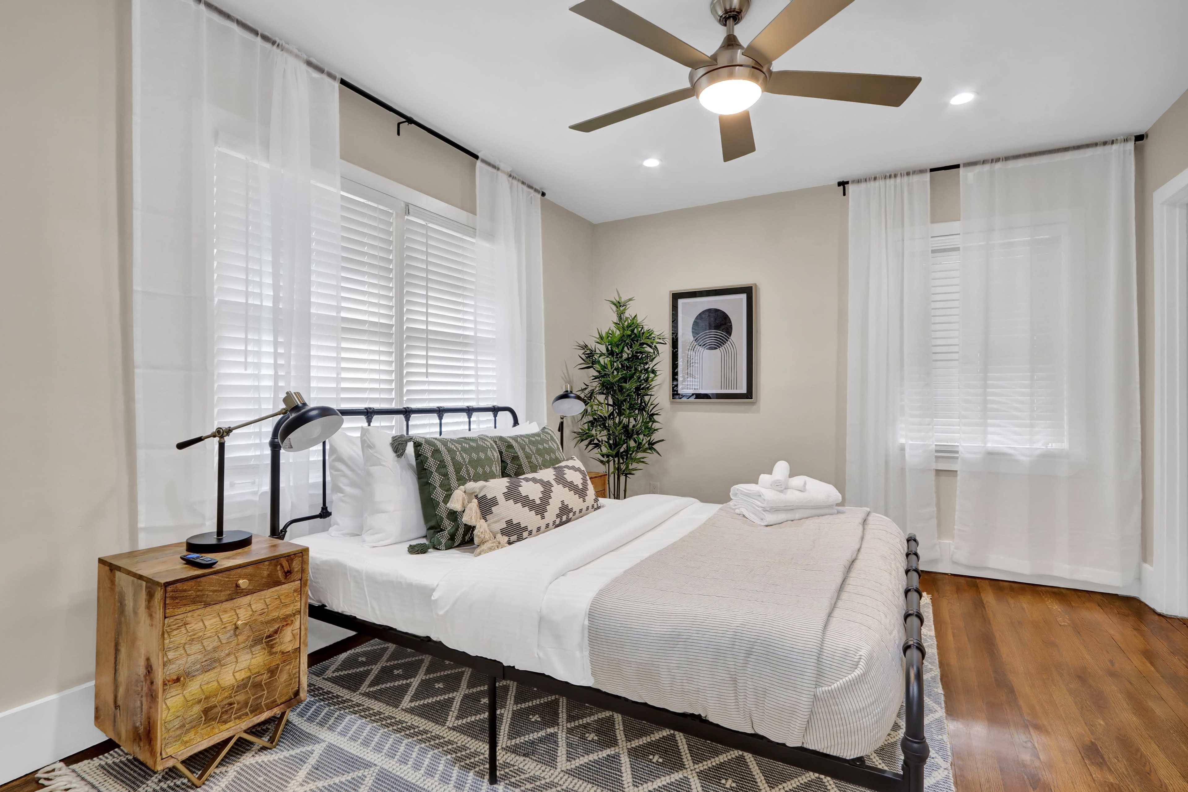 A neatly arranged bedroom featuring a bed with white linens, a wooden nightstand, and large windows with sheer curtains.