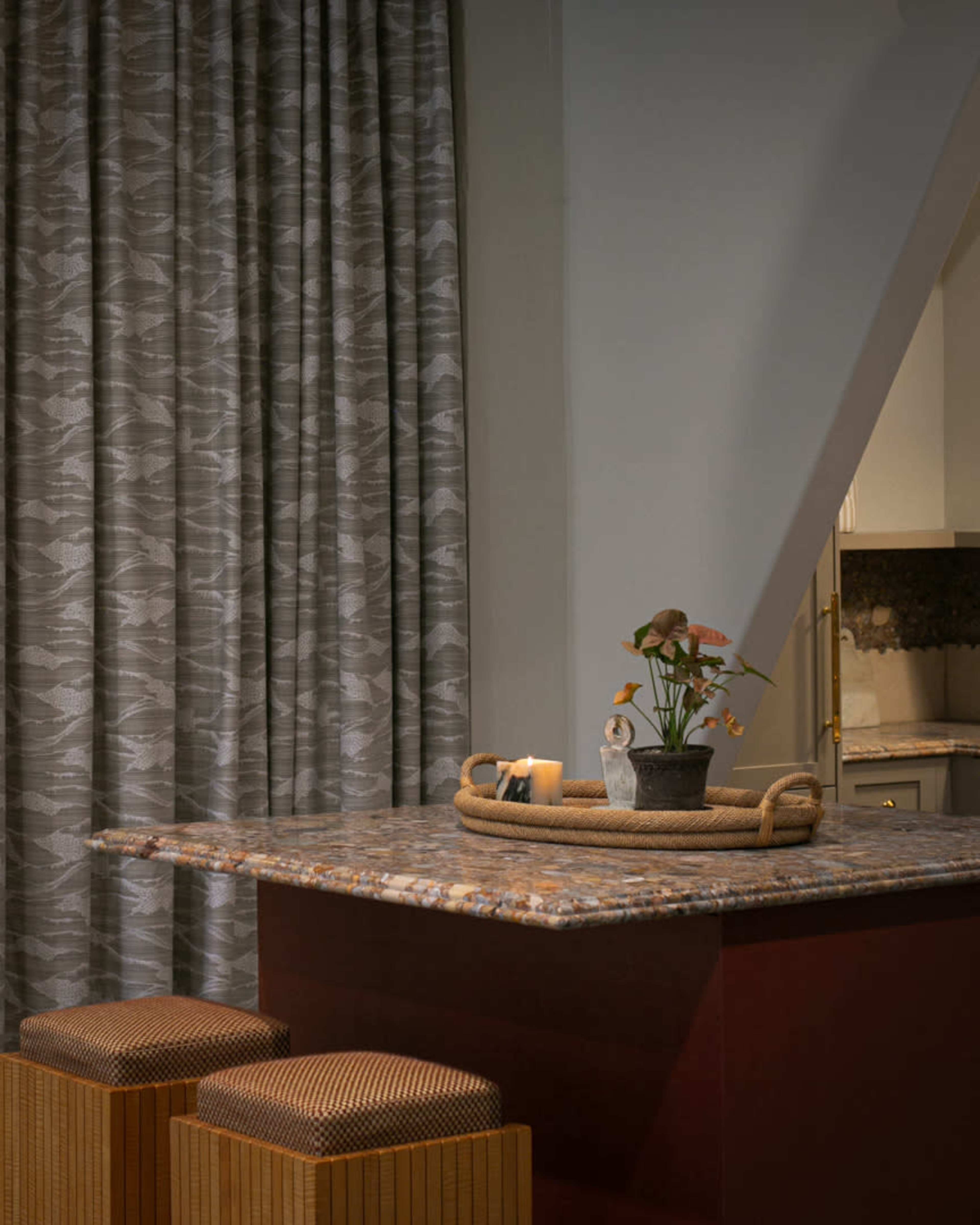A small kitchen area features a patterned curtain, a granite countertop with a potted plant and candle, and two woven stools.