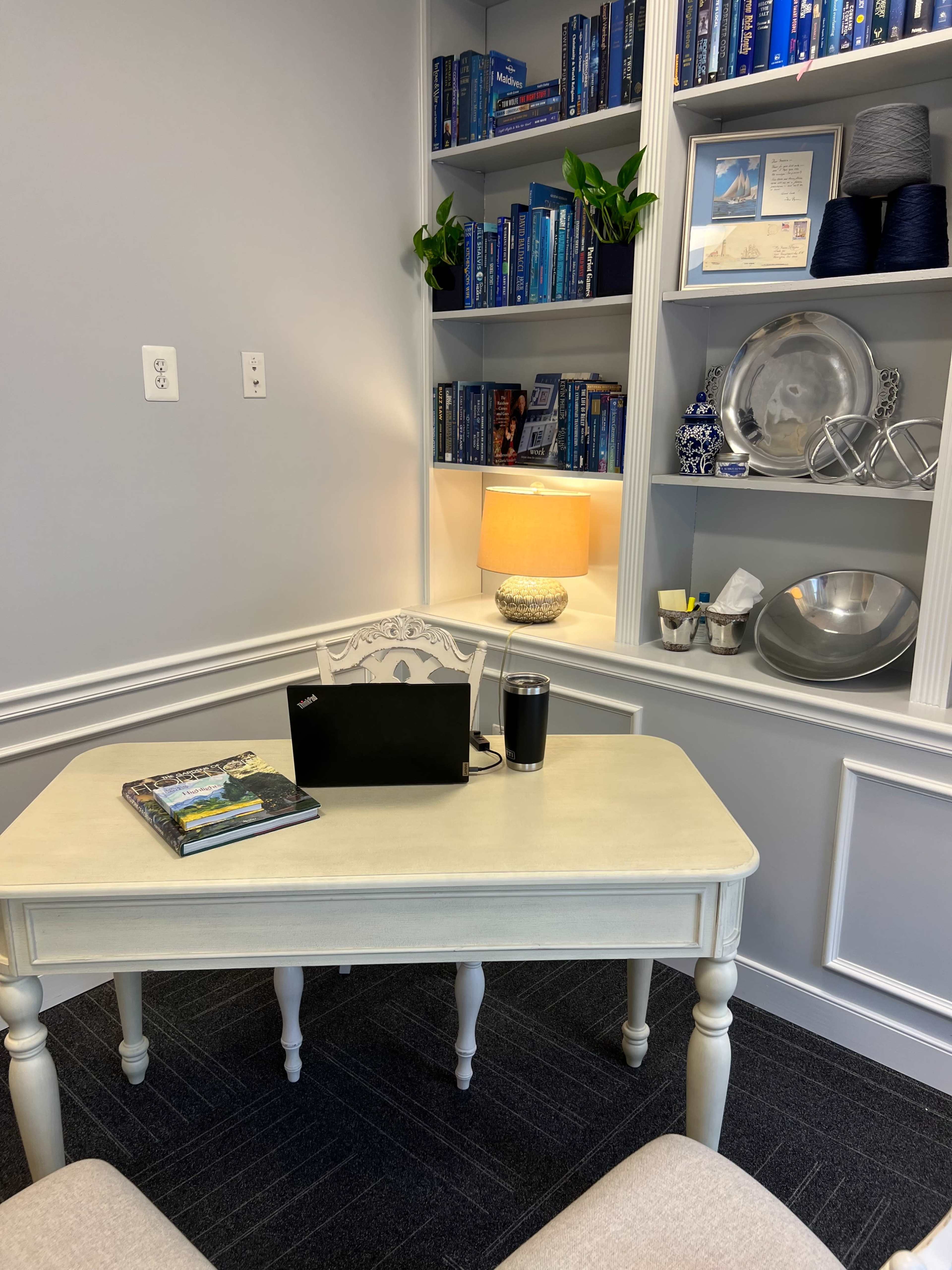 The image shows a small, well-organized office space with a light-colored desk, a laptop, a book on the desk, shelves filled with books and decorative items, and a lamp providing illumination.