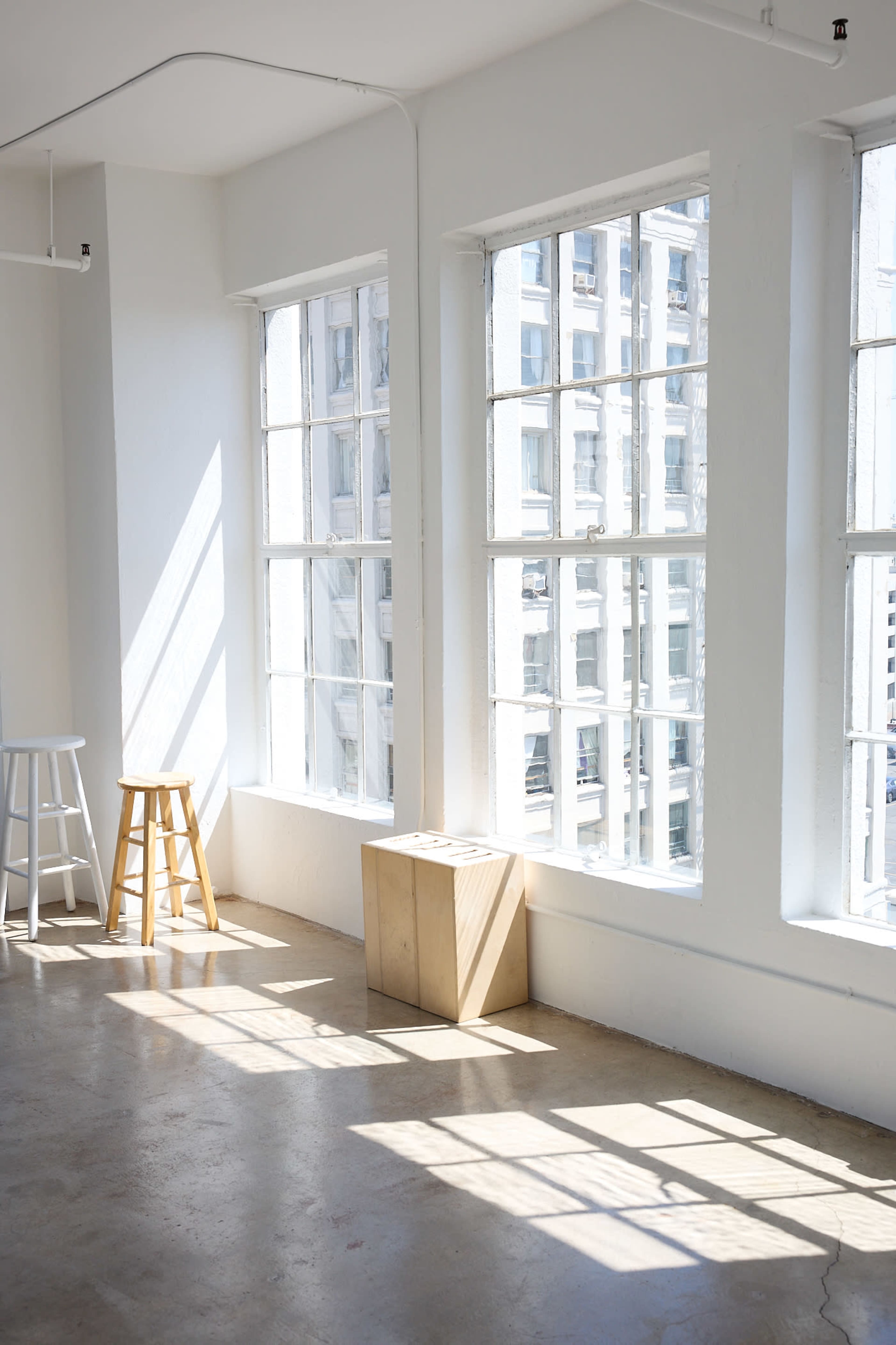A bright, airy room with large windows allowing sunlight to filter in, illuminating a wooden stool and a box near the windows.
