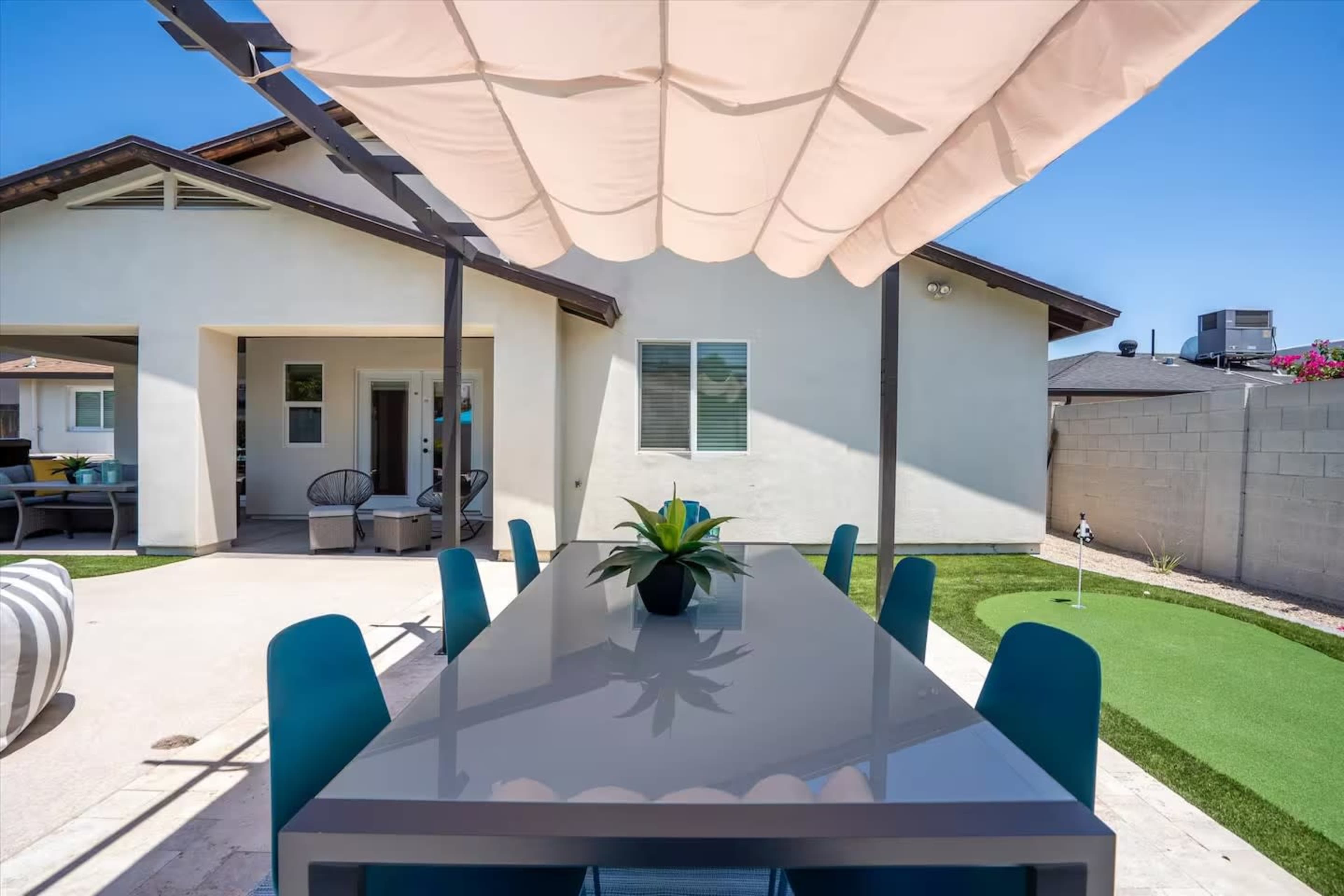 A long dining table with blue chairs is set up under a canopy in a backyard, adjacent to a house with a light-colored exterior.