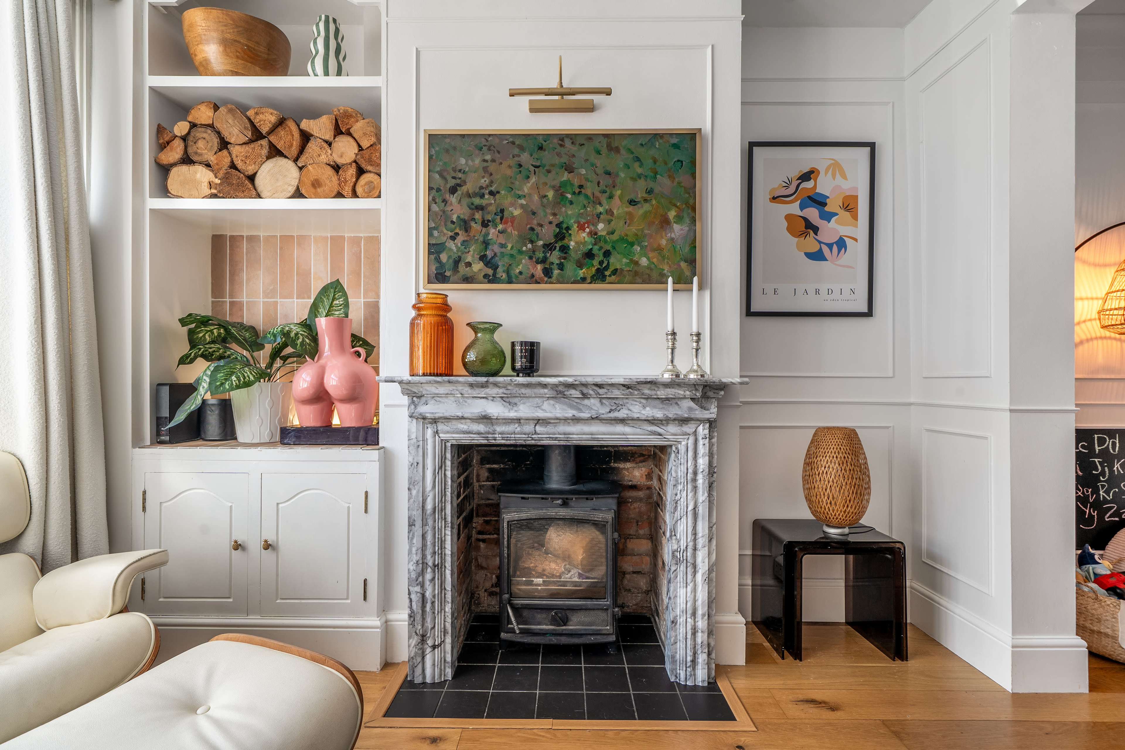 The image shows a cozy living room featuring a marble fireplace with a wood stove, surrounded by decorative artworks and a stacked log display.
