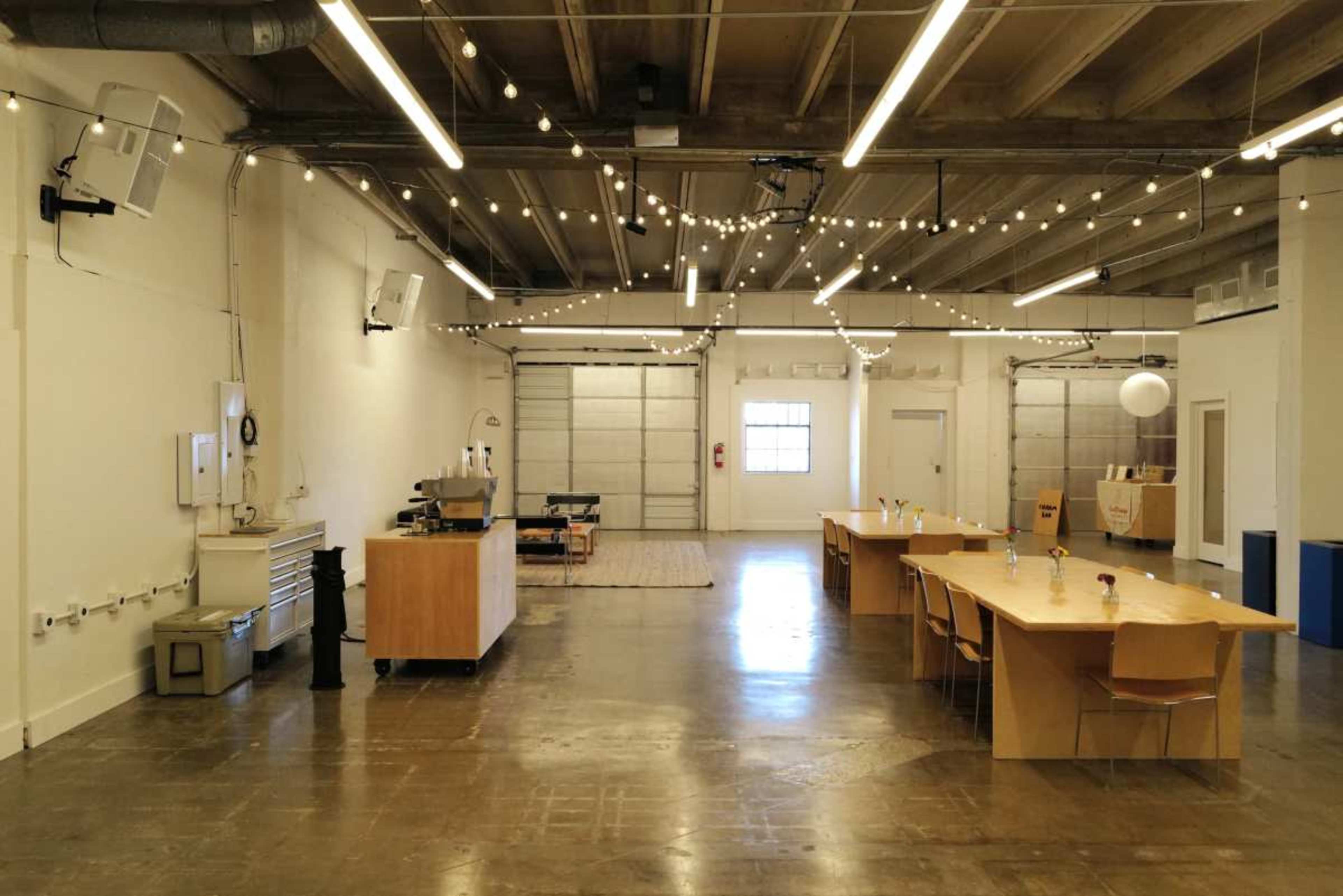 The image shows a spacious, well-lit room with a long table, chairs, a kitchen area, and decorative string lights hanging from the ceiling.