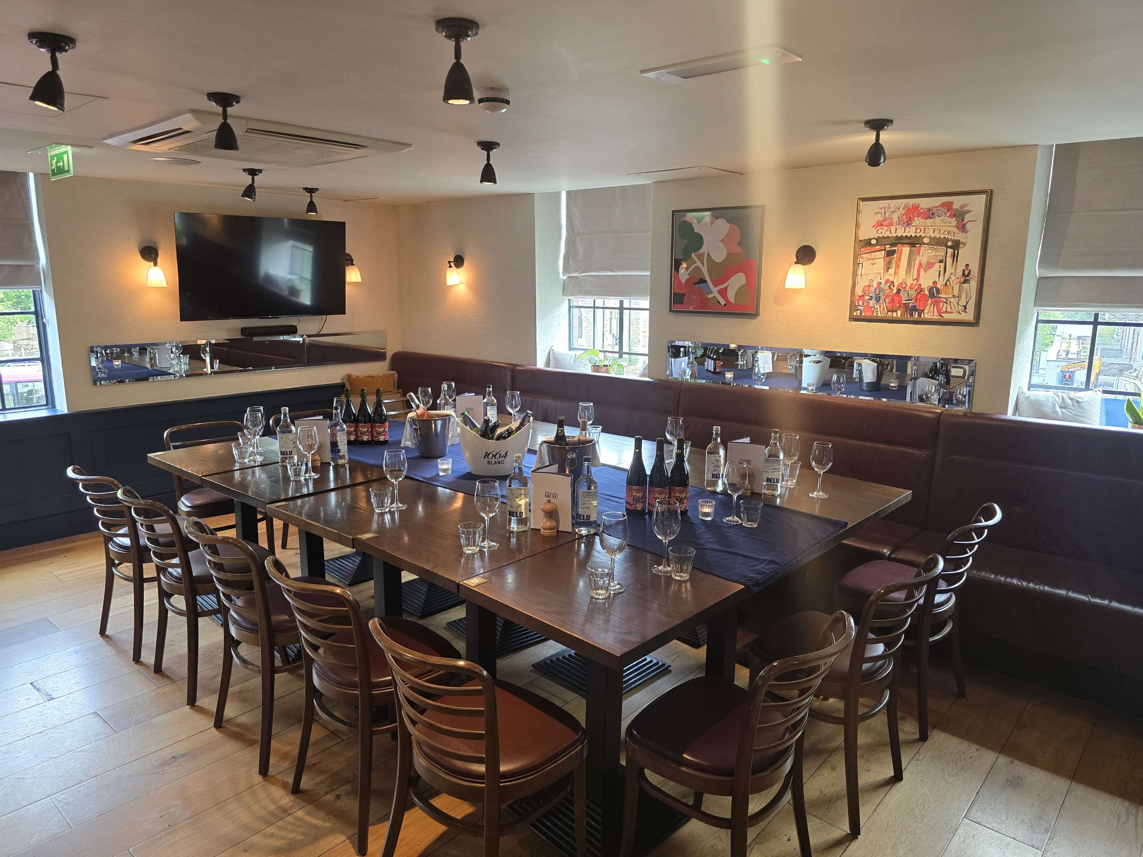 A spacious dining room features a long table set with glasses and bottles, surrounded by wooden chairs and framed artwork on the walls.