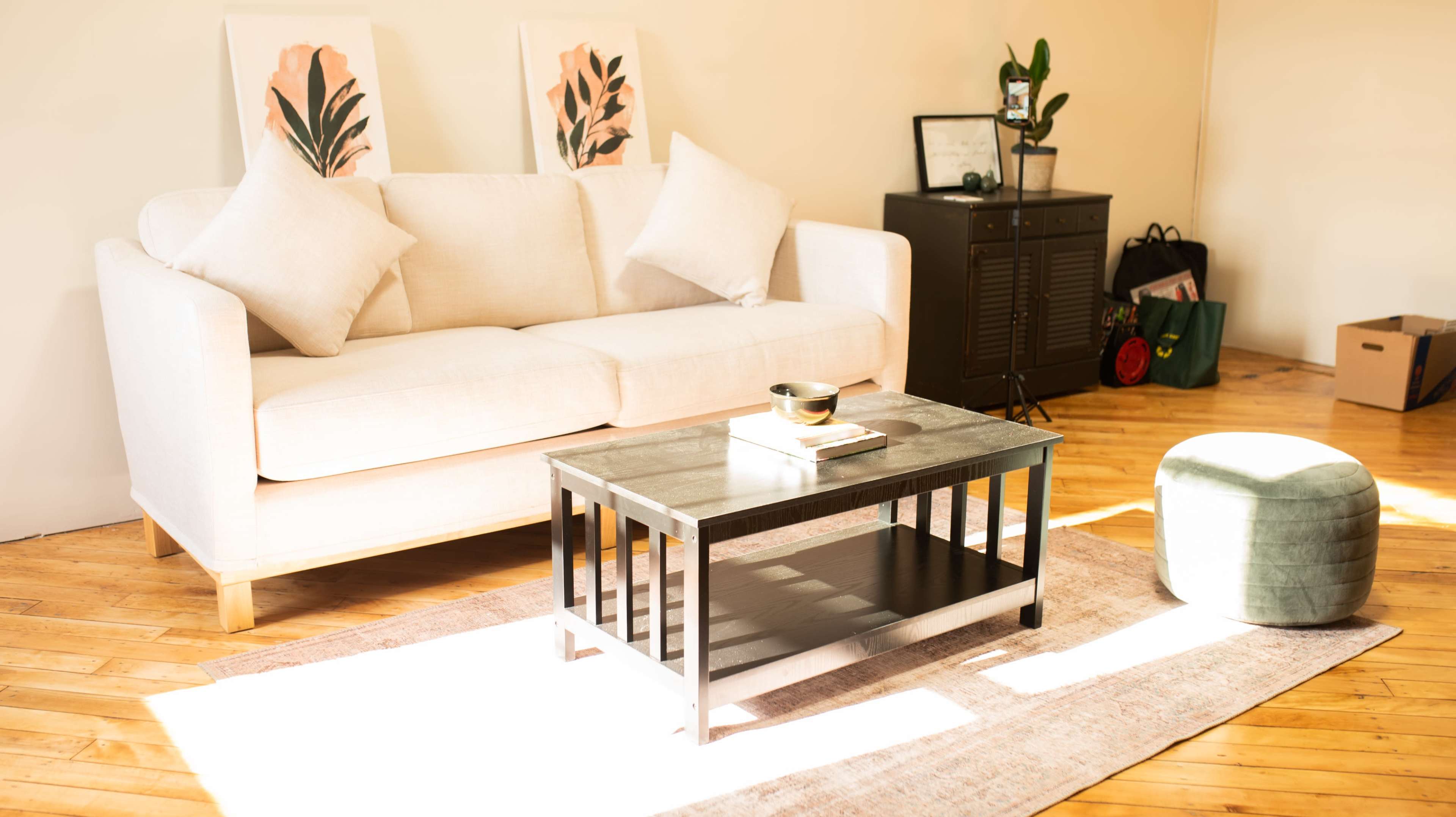A light-filled living room features a beige sofa with two cushions, a black coffee table, and a round green ottoman, set on a wooden floor with a rug underneath.