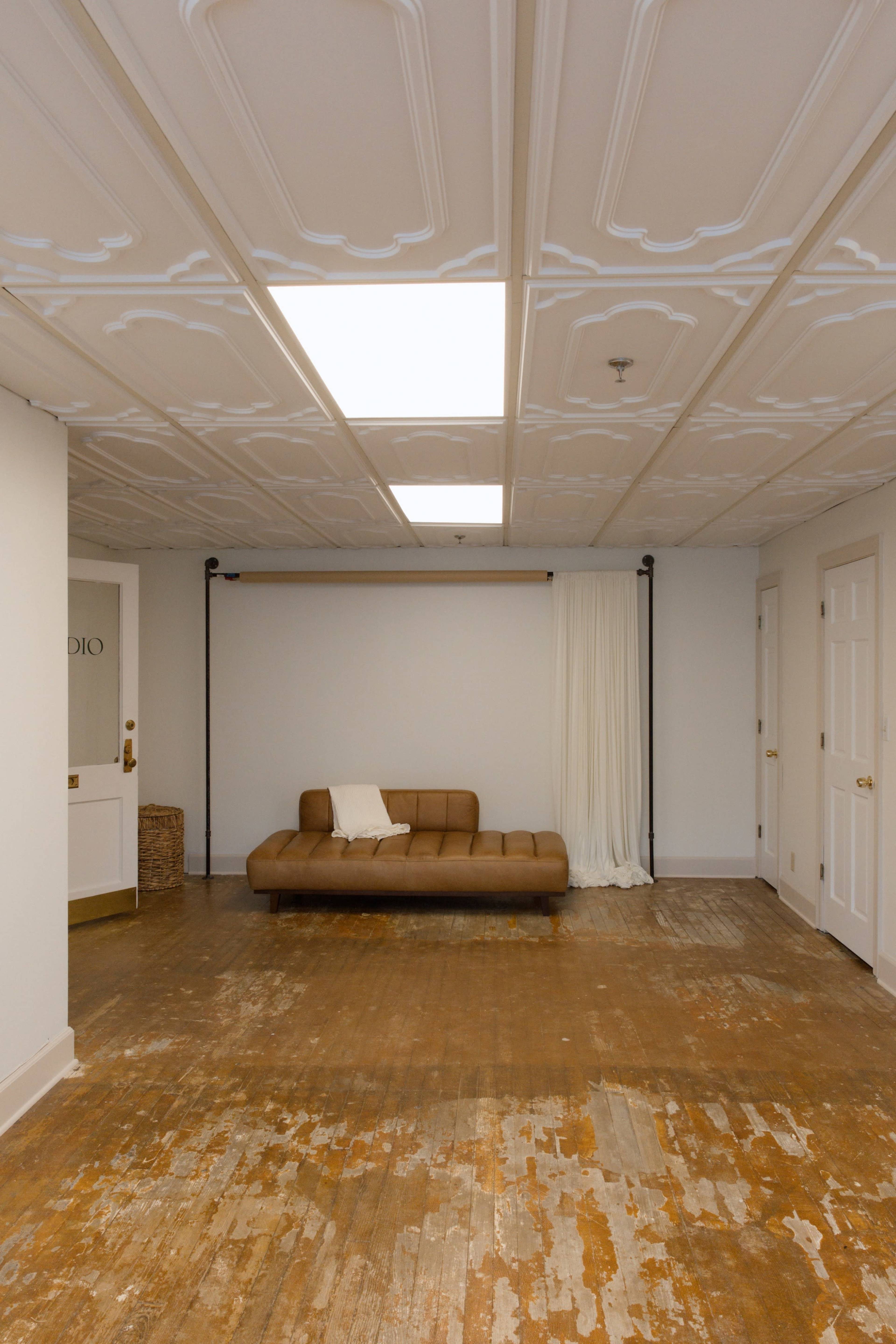 The image shows a spacious room with a brown sofa, wooden floor showing signs of wear, and white walls with a draped curtain and a light fixture on the ceiling.