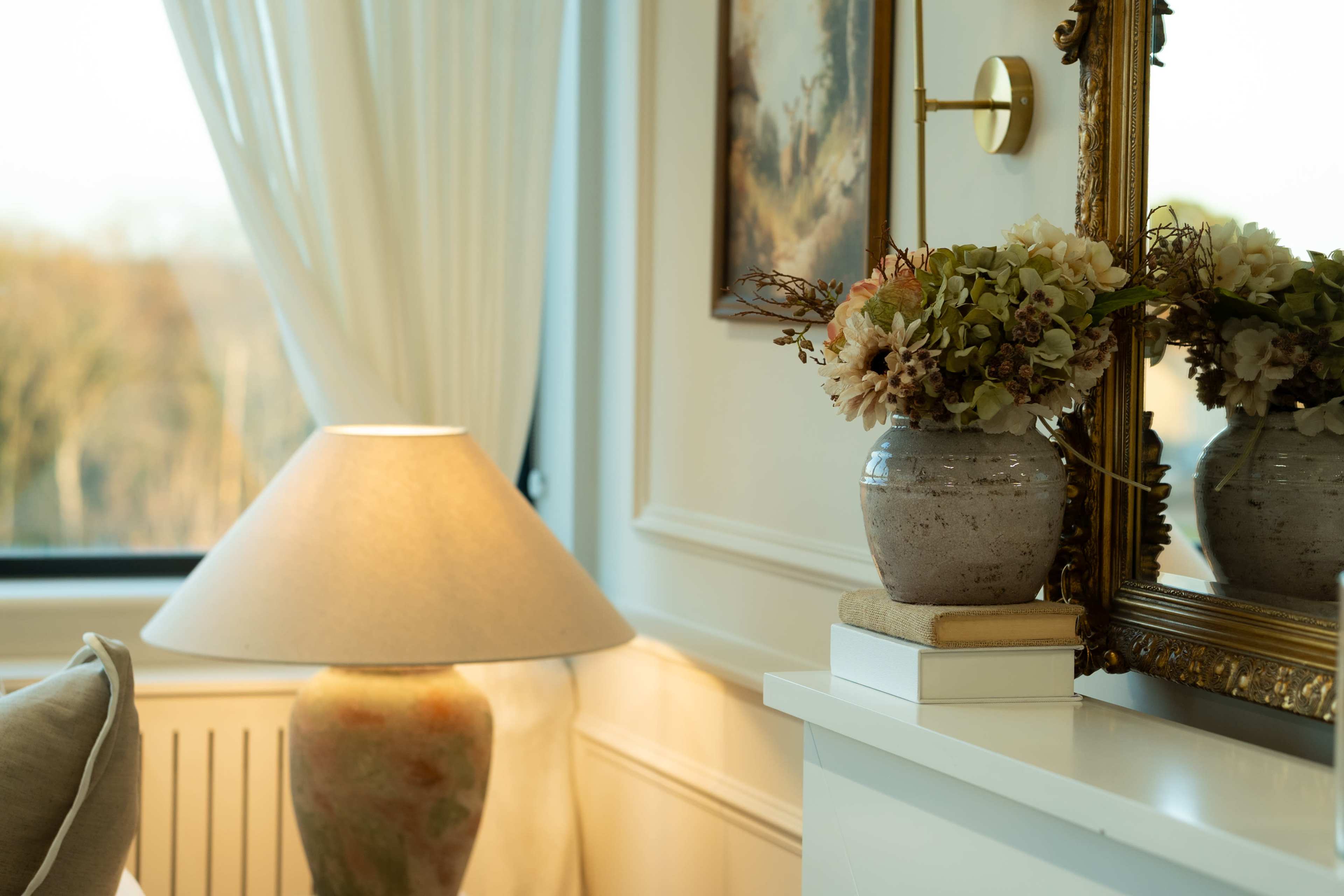 The image features a cozy interior scene with a lamp, a vase of flowers, and a decorative mirror reflecting the room’s ambiance.