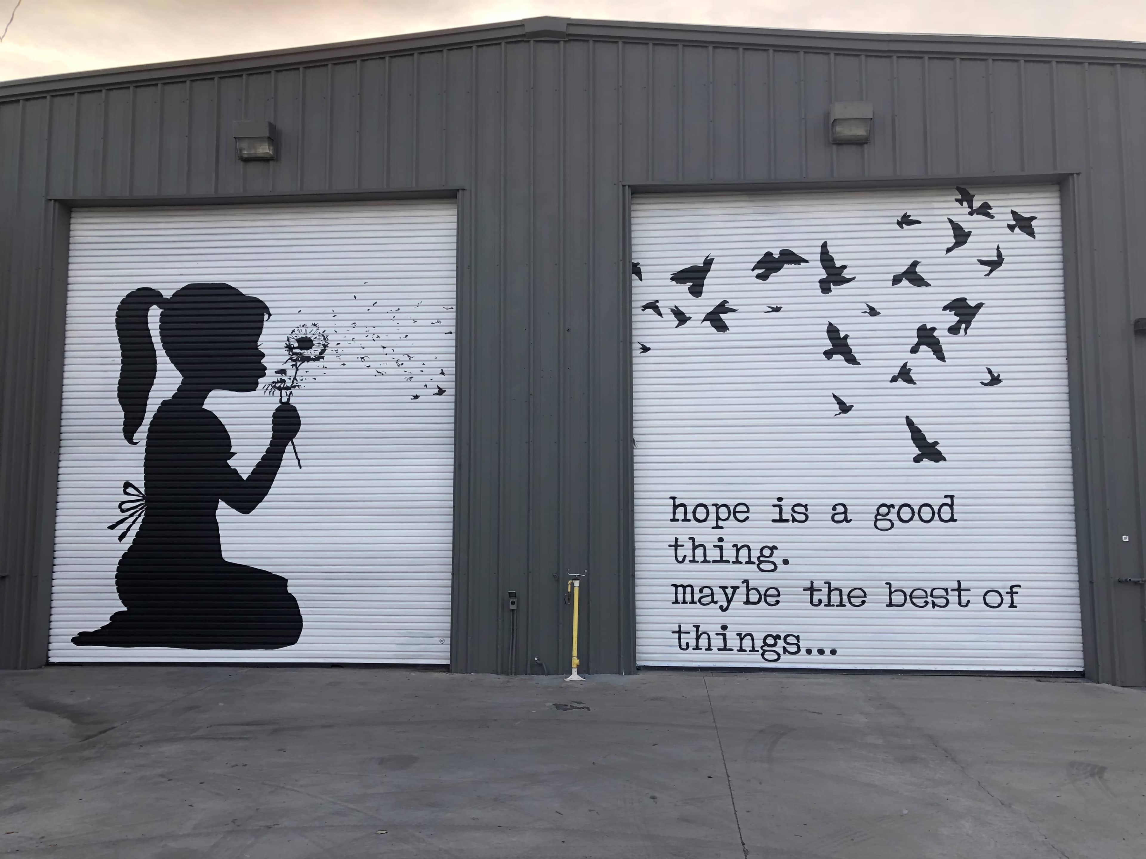 The image shows two garage doors featuring a black silhouette of a girl kneeling and blowing a dandelion, accompanied by flying birds and text that reads, "hope is a good thing. maybe the best of things..."