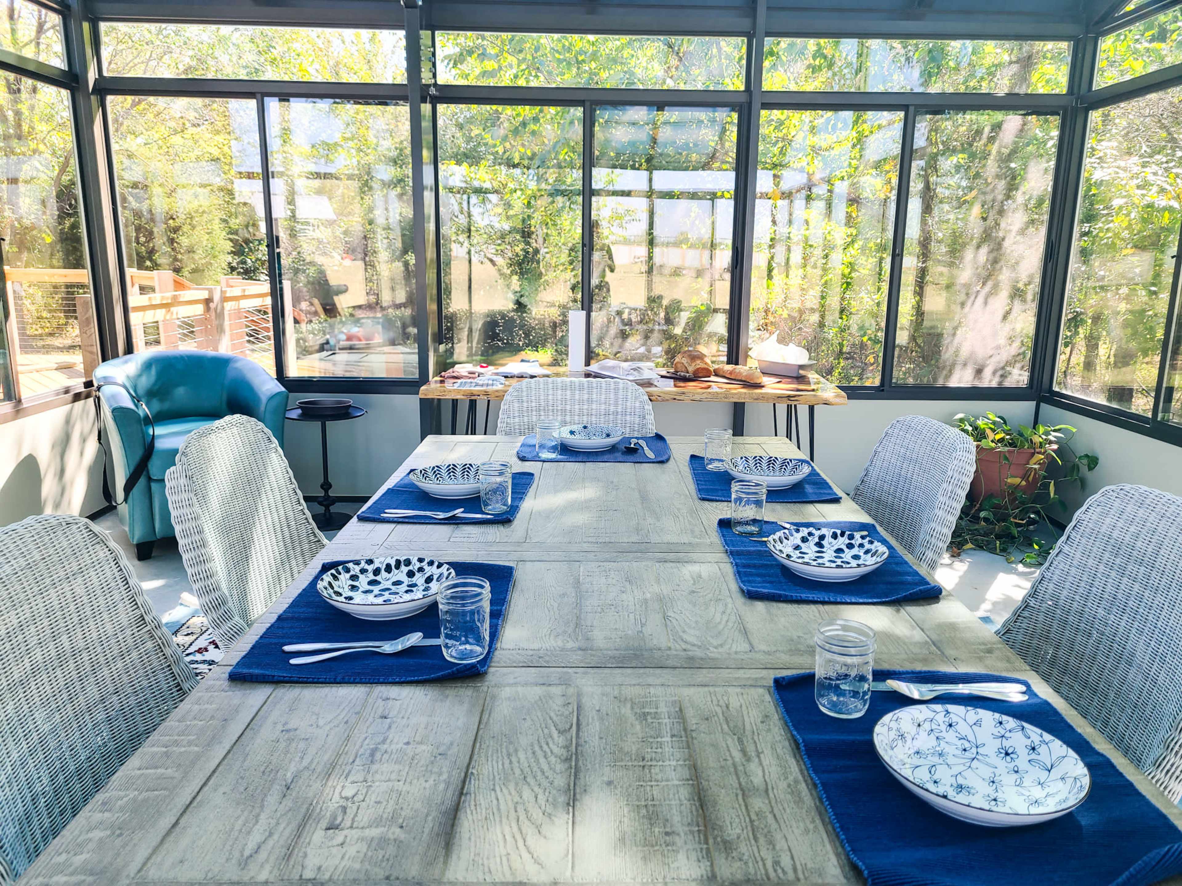 A dining table is set with plates and utensils in a sunlit glassed-in porch surrounded by greenery.