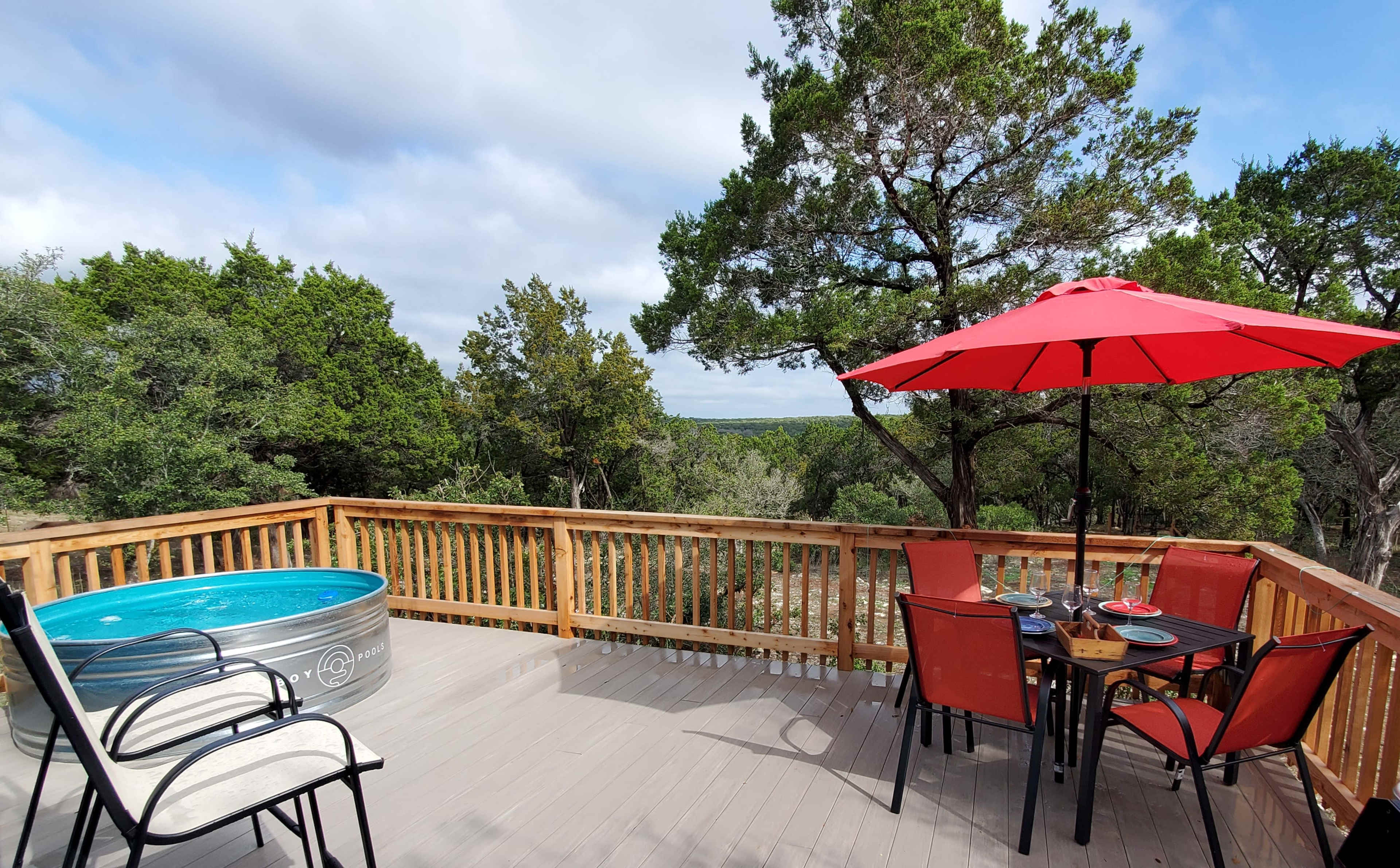 A wooden deck featuring a round metal swimming pool, a red umbrella over a table, and chairs surrounded by trees and a distant landscape.