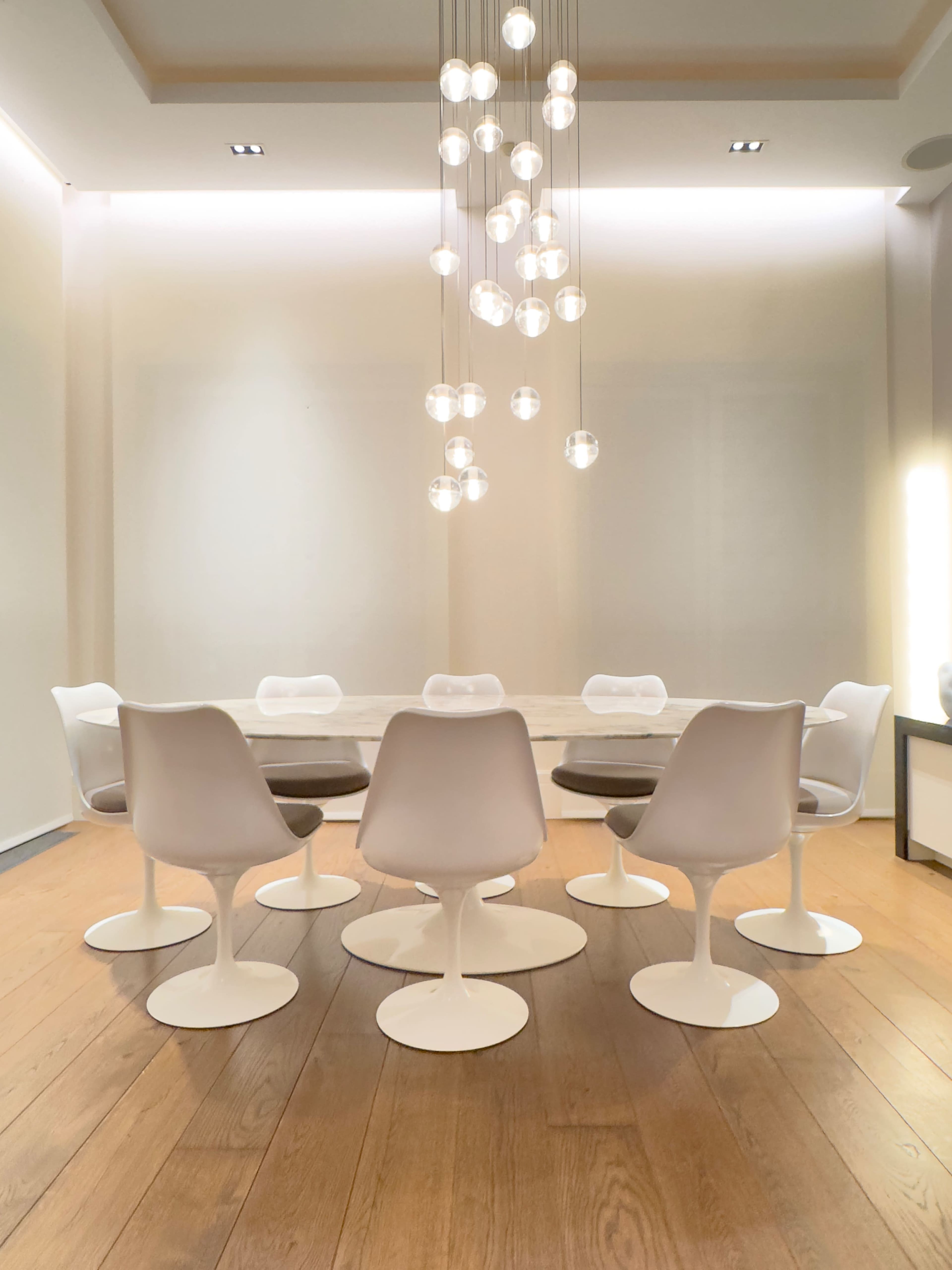 A modern conference room features a round table surrounded by white chairs and a cluster of pendant lights above.