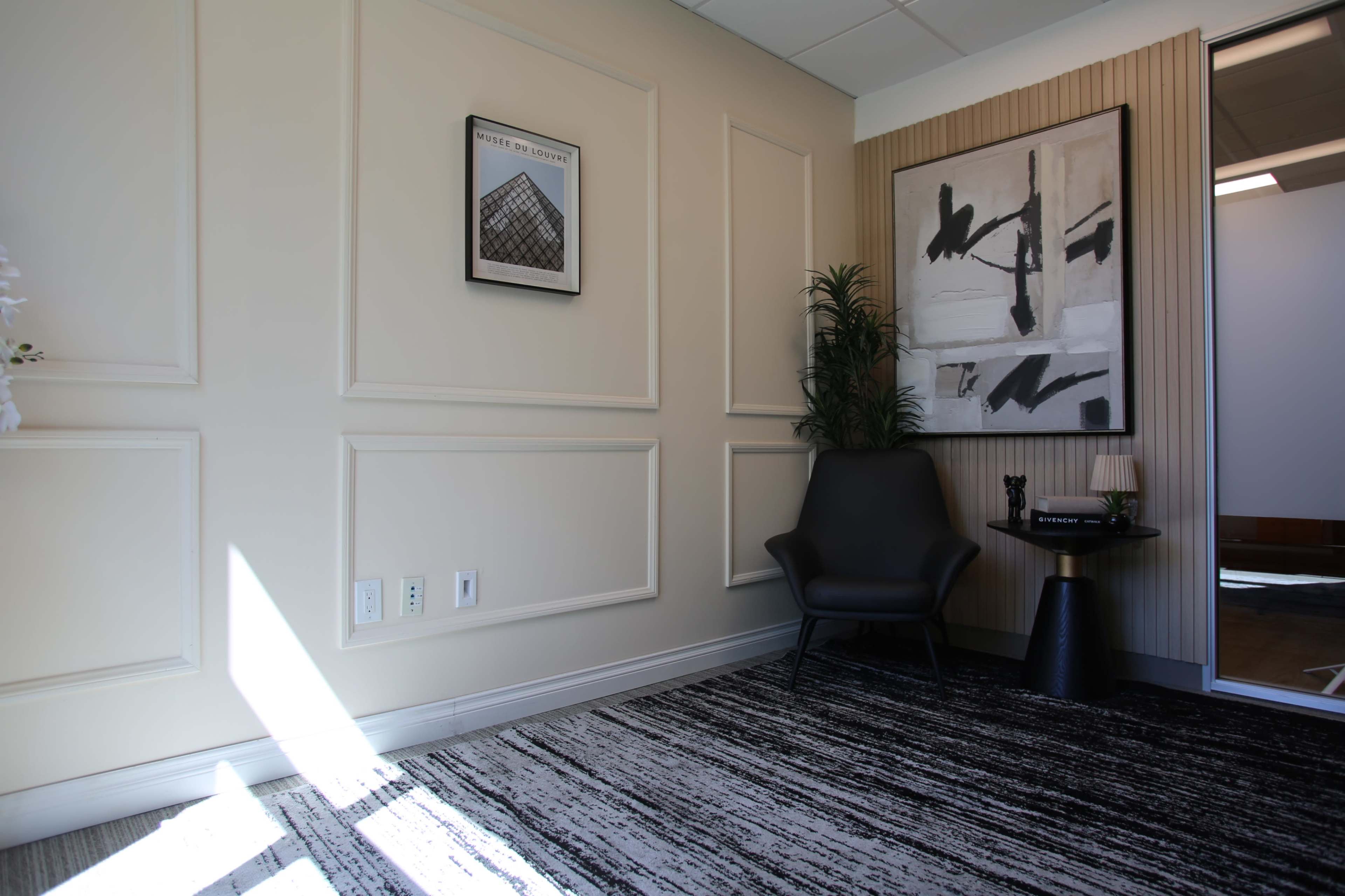 The image shows a modern office corner featuring a black chair, a small round table, a decorative artwork on the wall, and natural light illuminating the space.